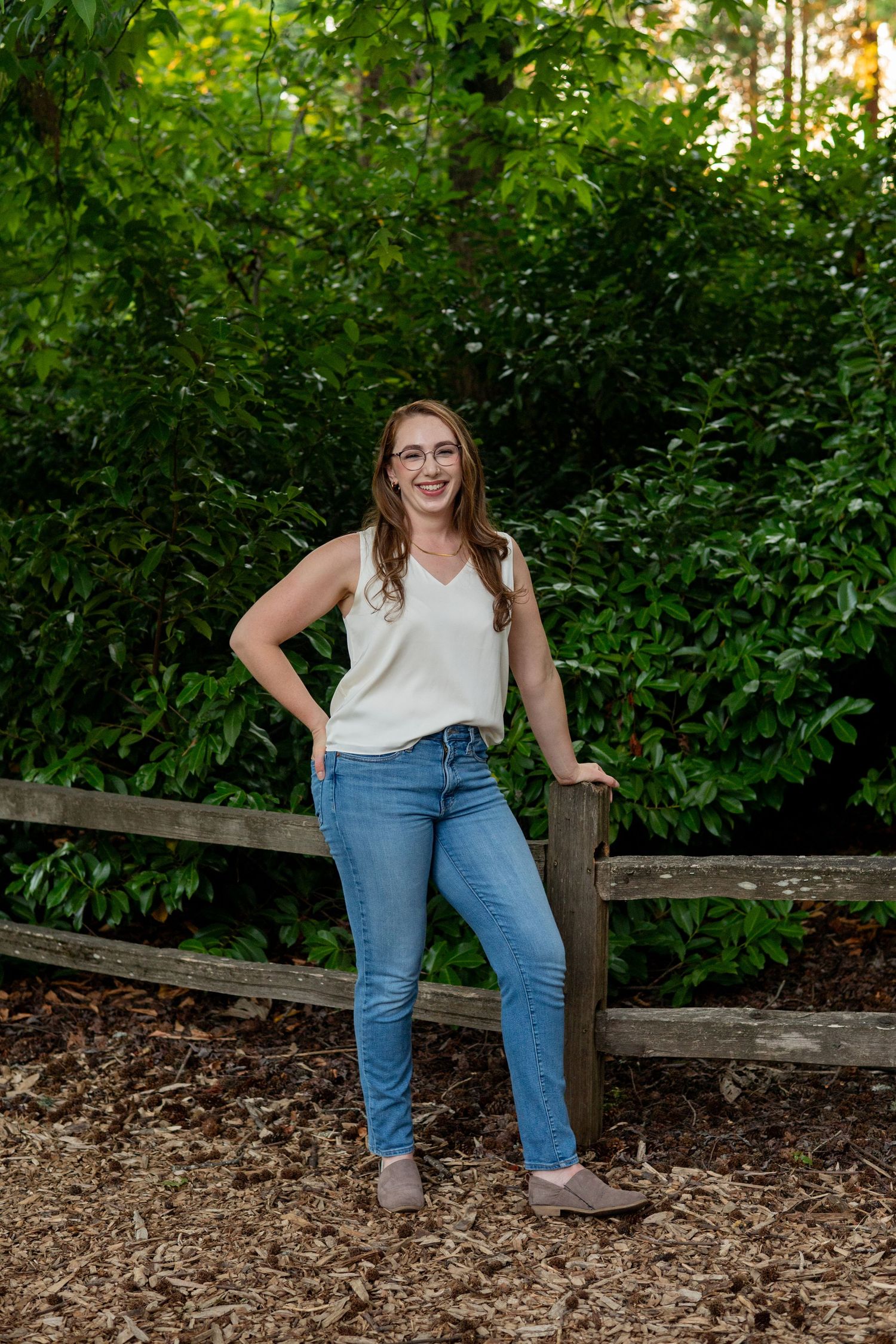 A person in a white top and blue jeans poses casually on a rustic wooden fence surrounded by lush green foliage.