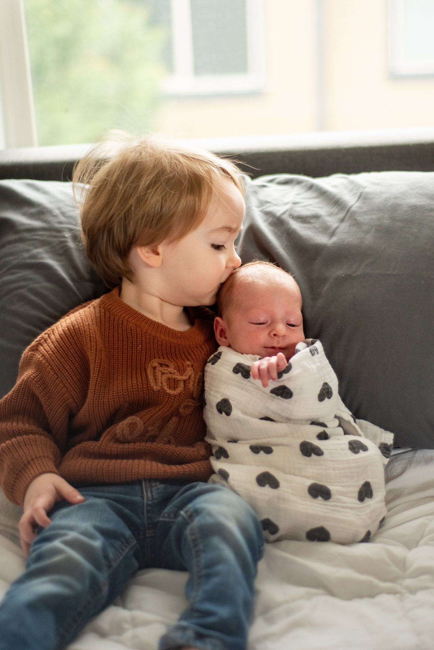 A toddler in an orange sweater gently kisses a swaddled newborn on a gray couch.
