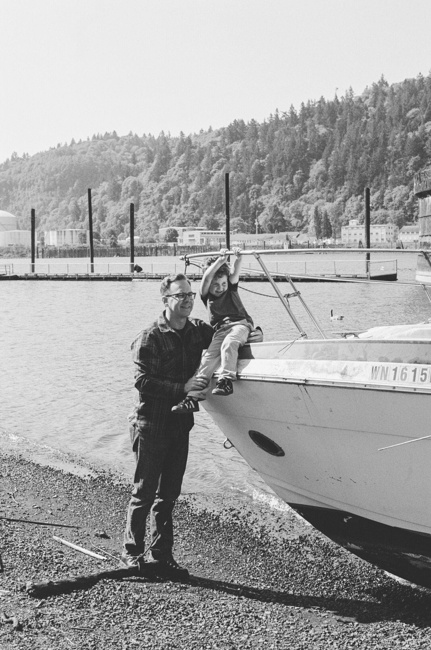 A black and white photo shows a docked boat at a marina with mountains and forest in the background.