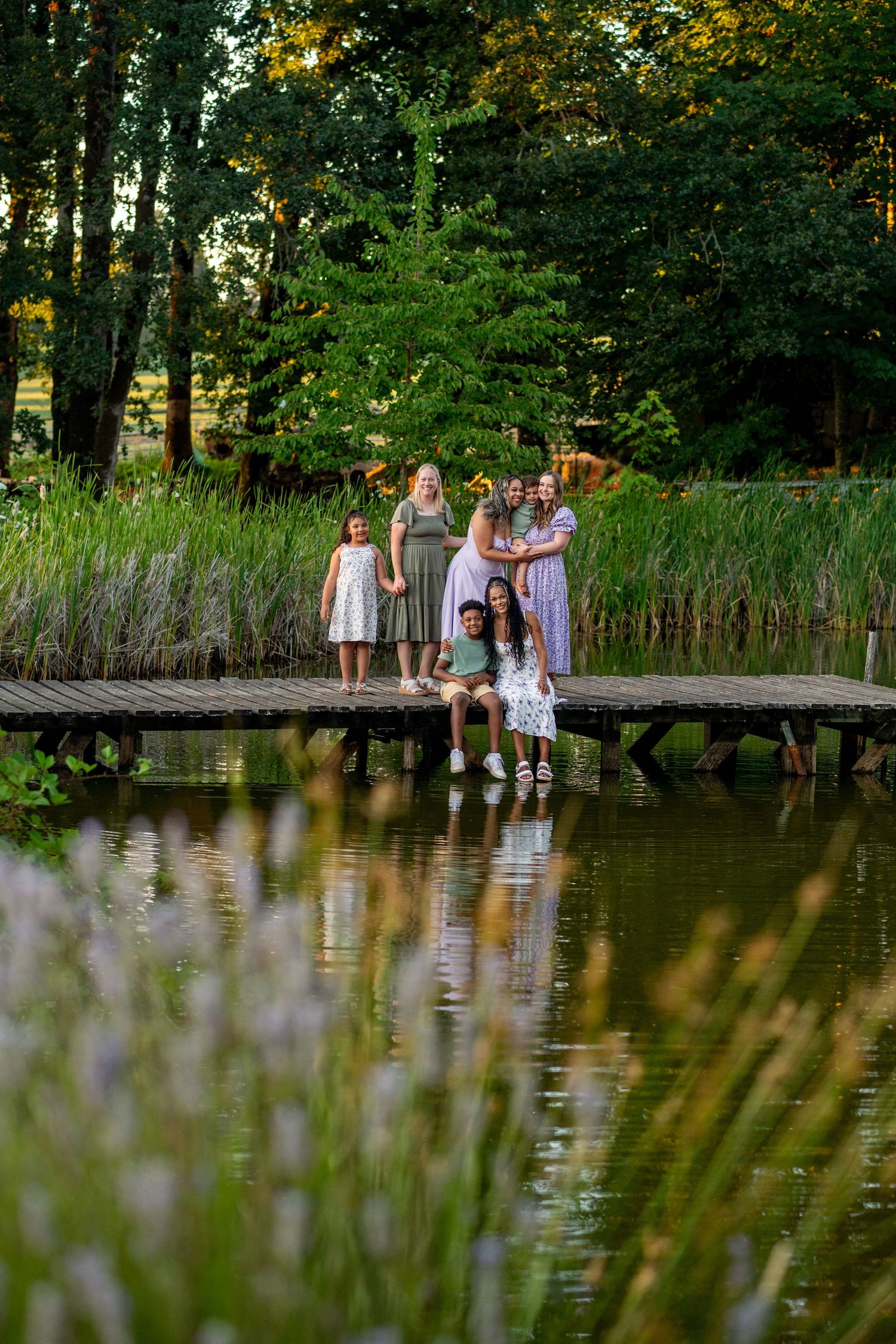 Family in light summer clothing poses on a wooden dock by a tranquil pond with cattails and trees in the background.