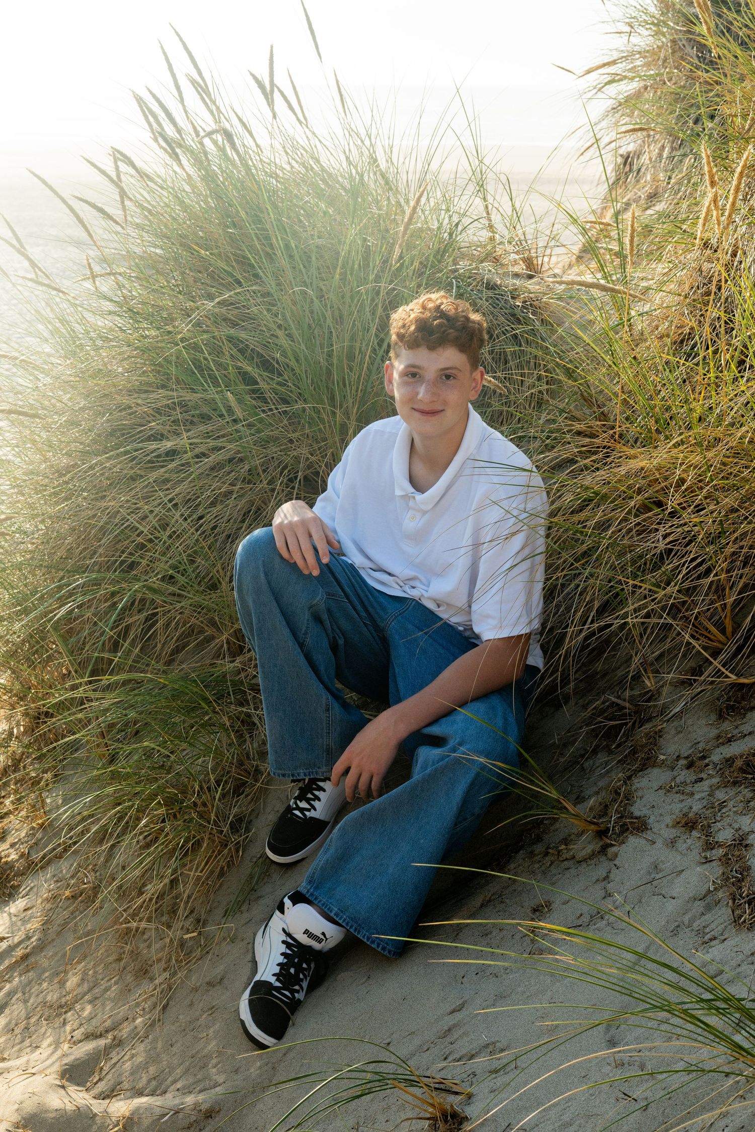 Young person in white shirt and blue jeans sitting on beach dunes with sea grass.