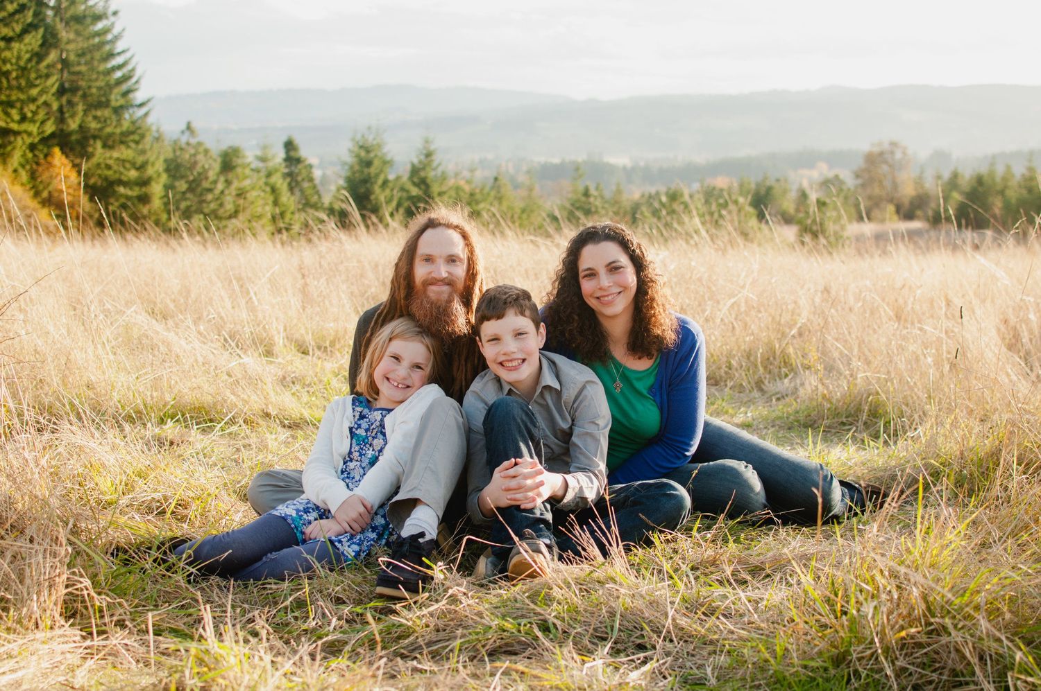 A casual outdoor family portrait in a golden wheat field with mountains in the background.