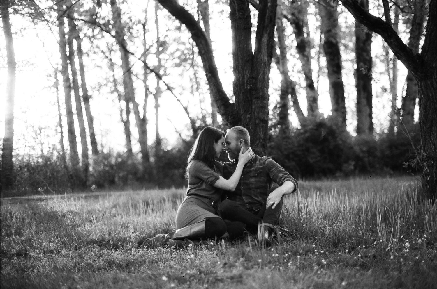 A black and white photo of two people embracing on grassy ground with sunlit trees in the background.