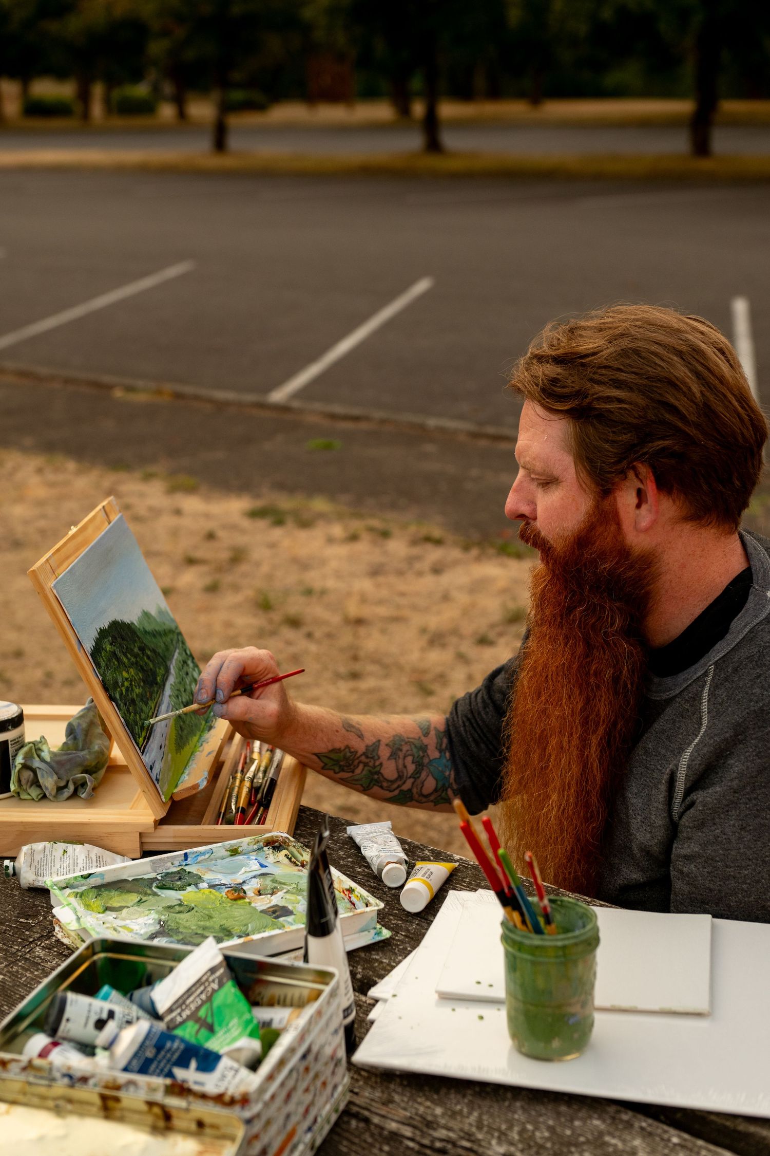 Artist with long red beard works intently on painting at an outdoor table with art supplies and paper.