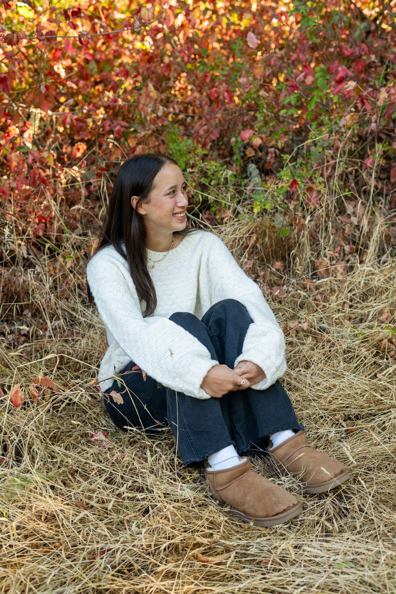 A person wearing a white sweater and jeans sits on autumn grass against a backdrop of colorful fall foliage.