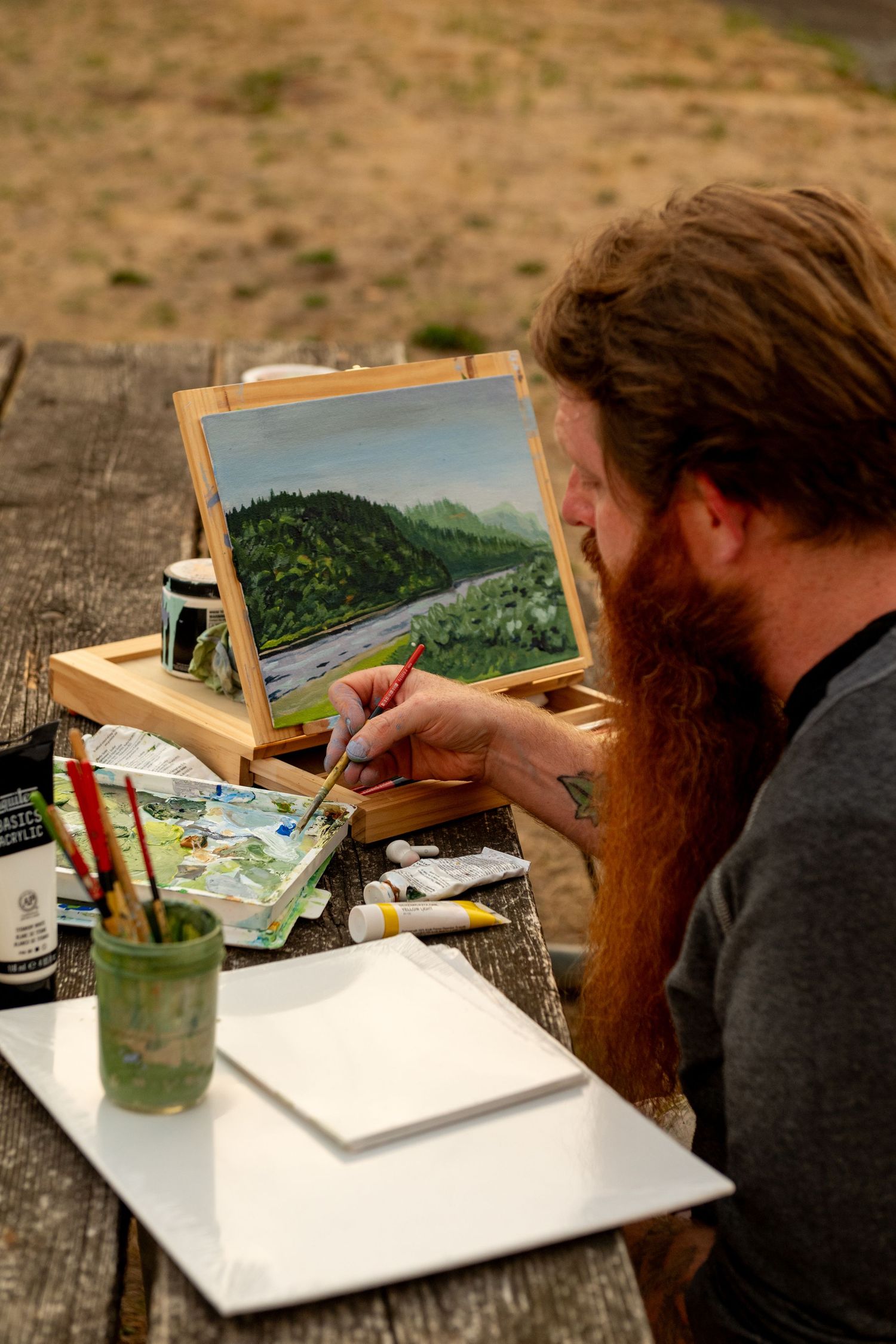 Artist sits at wooden table outdoors painting a watercolor landscape scene with mountains and forest.