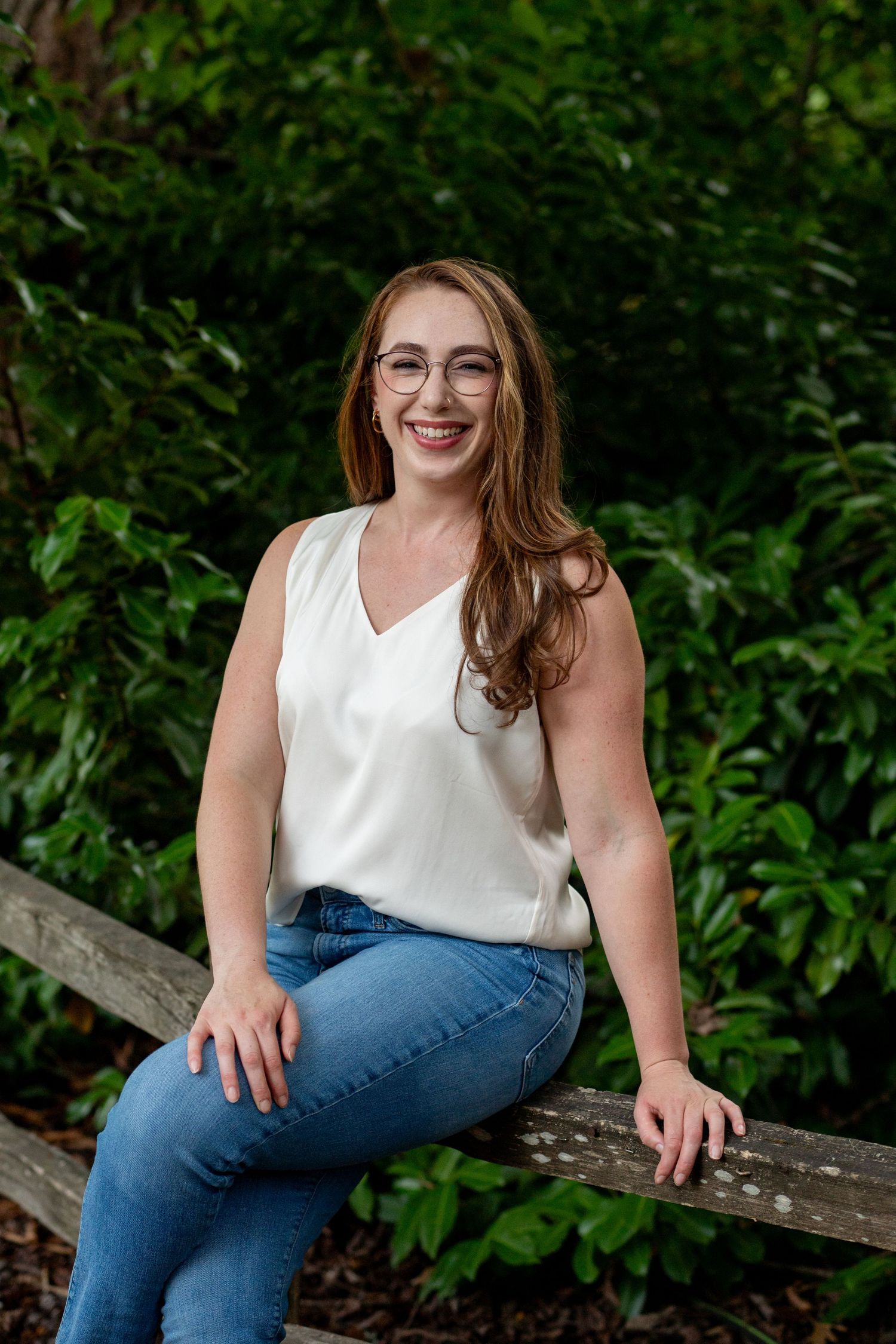 A person in a white sleeveless top and blue jeans sits casually on a wooden bench surrounded by lush greenery.