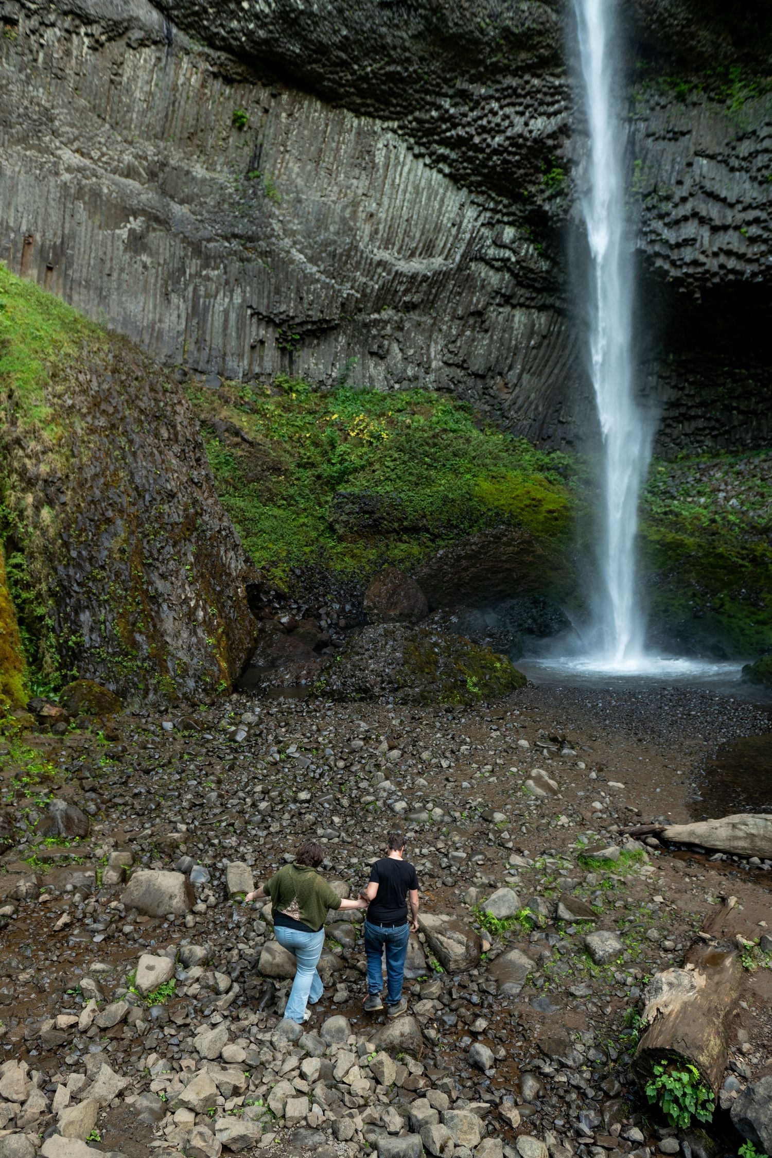 A dramatic waterfall cascades down a mossy cliff face into a rocky basin surrounded by forest terrain.
