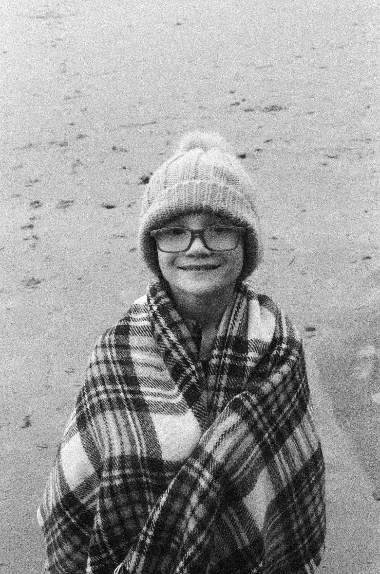 A person wrapped in plaid blanket wears winter hat and glasses on beach.
