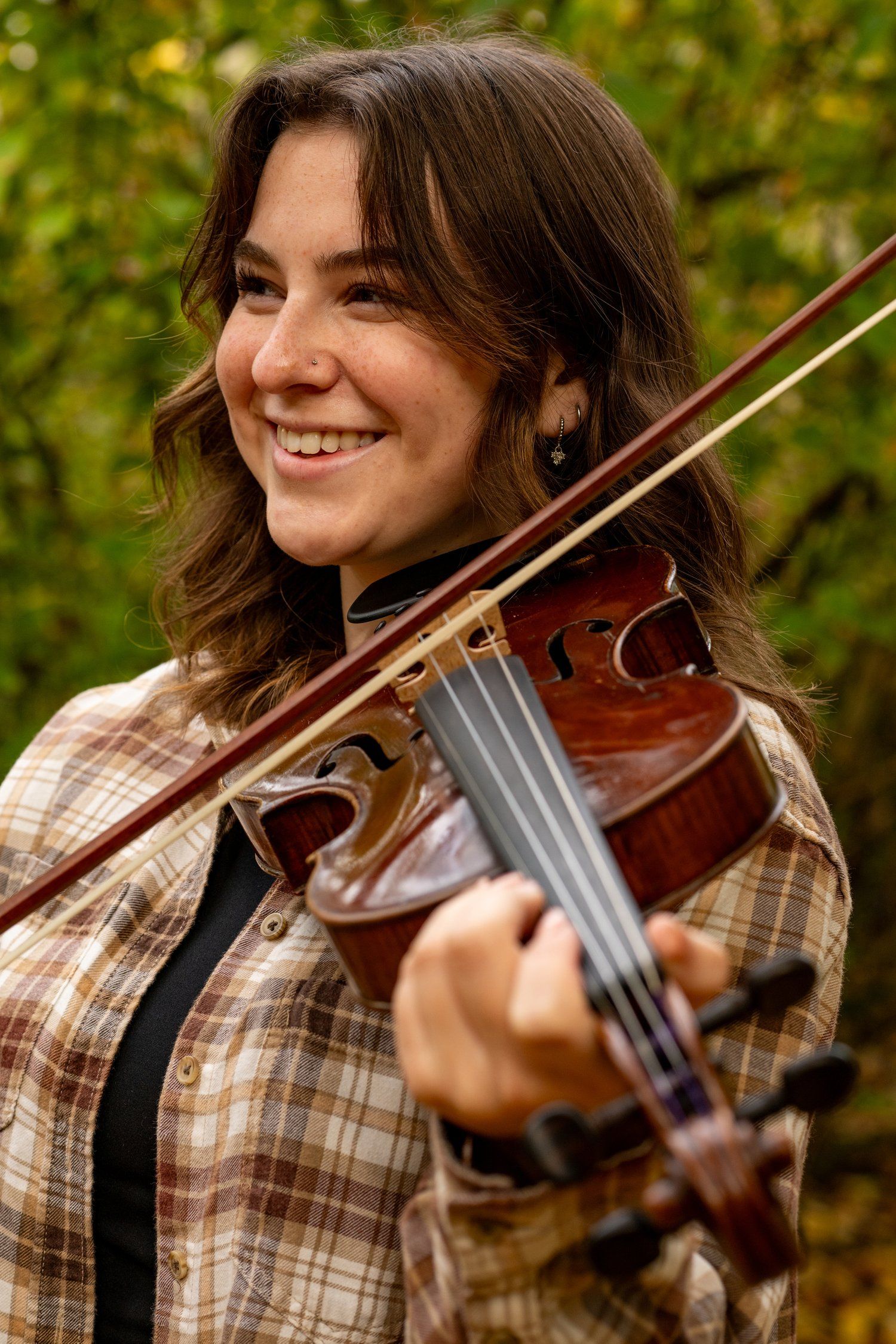 A musician plays violin outdoors while wearing a plaid shirt with autumn foliage in background.