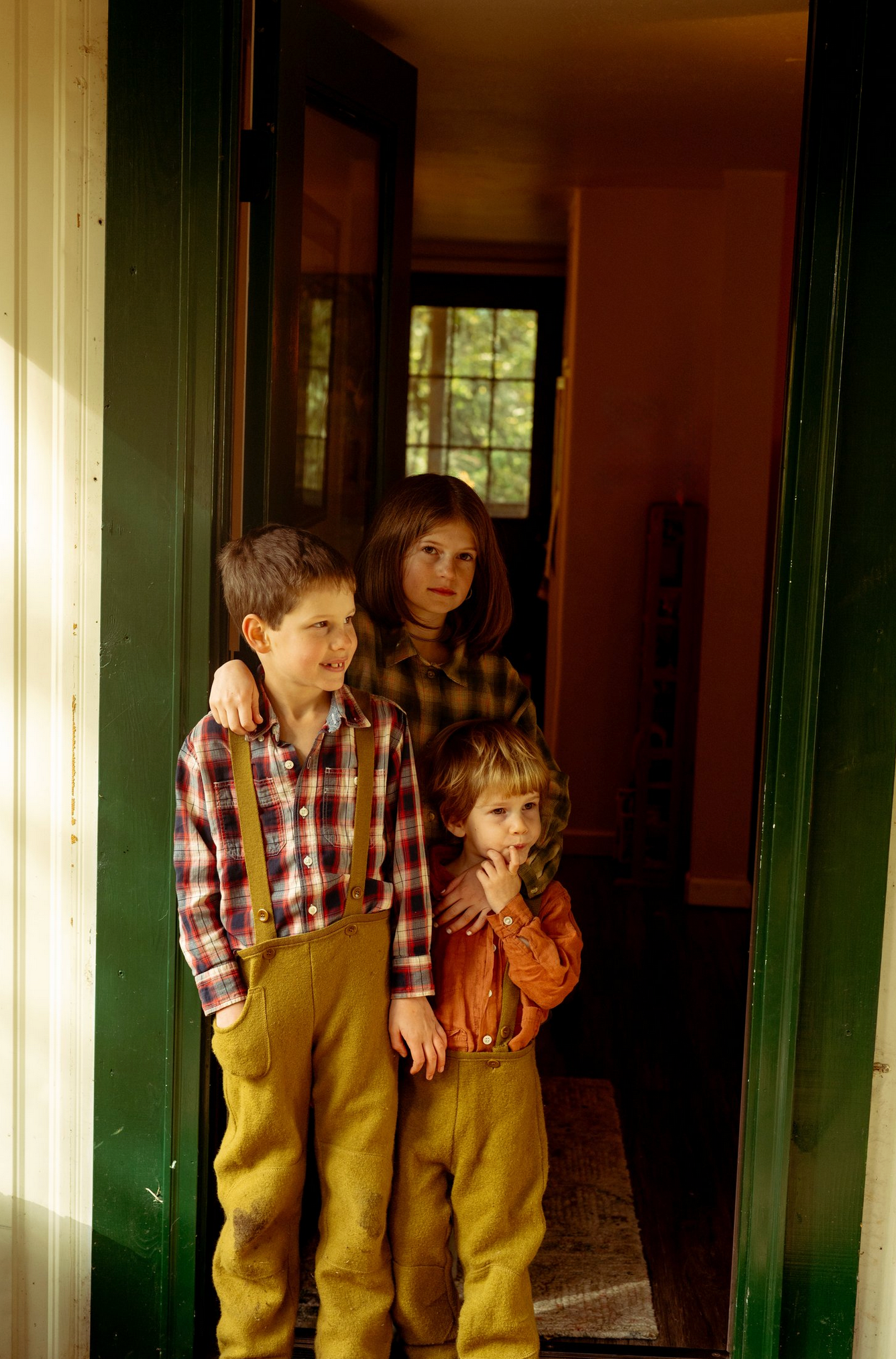 Children in plaid shirts and overalls pose playfully by vintage green doorway.
