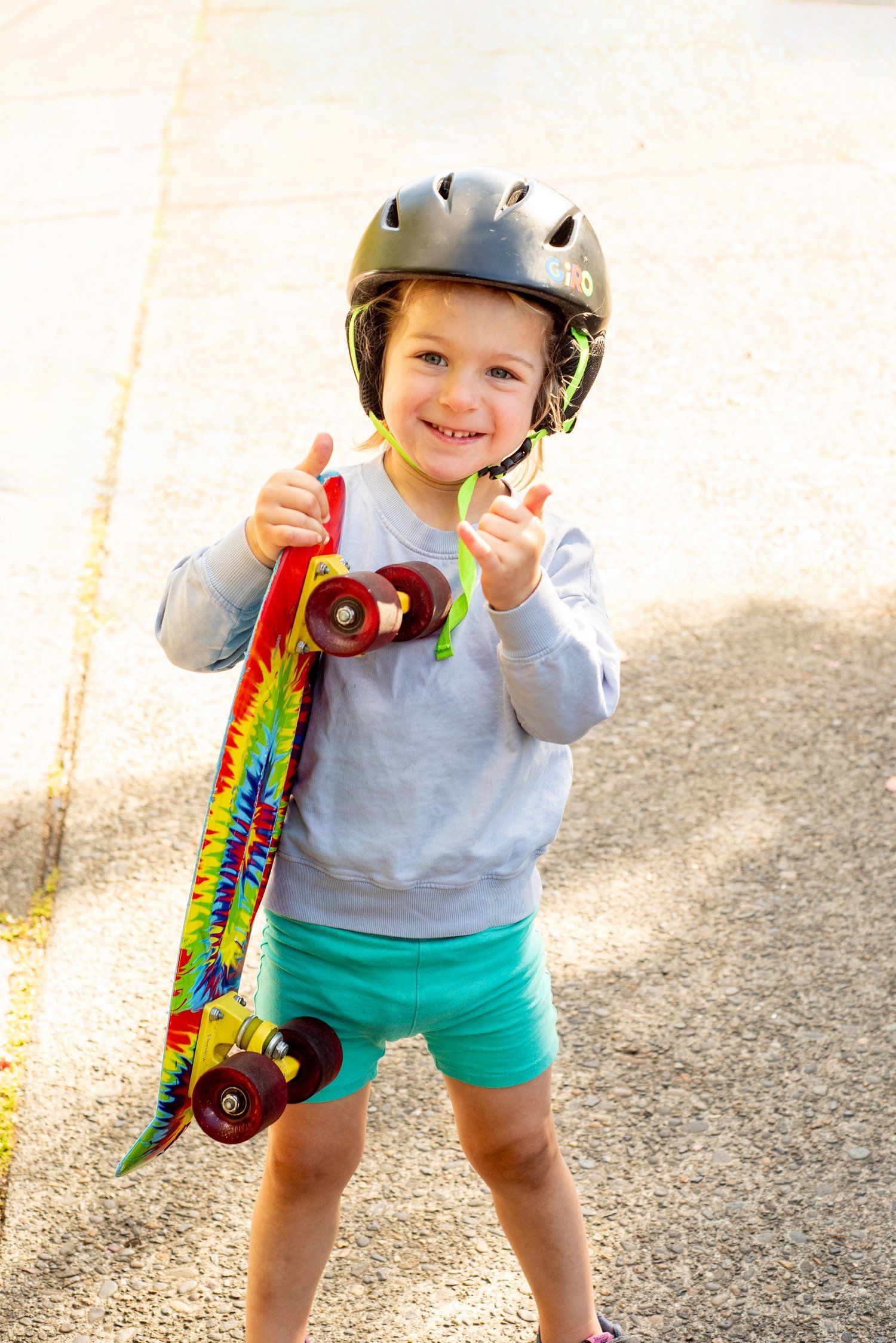 Child wearing helmet and turquoise shorts poses with colorful skateboard in sunlit setting.