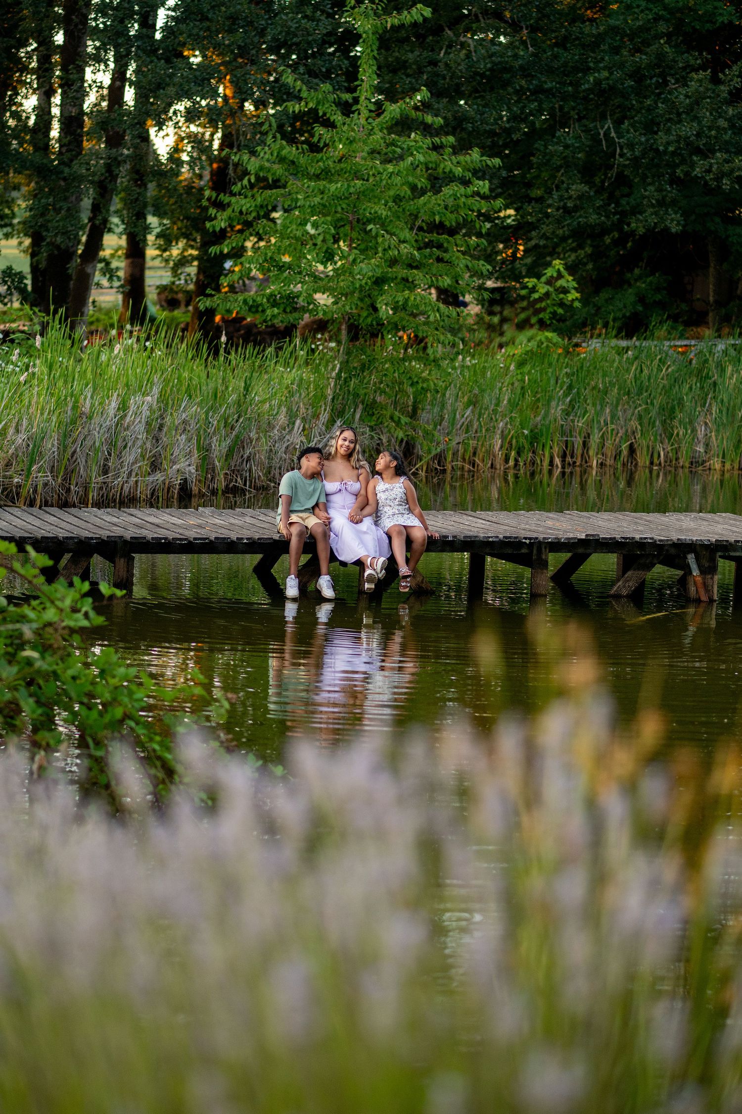 A peaceful lakeside scene shows three people sitting together on a wooden dock surrounded by reeds and green foliage.