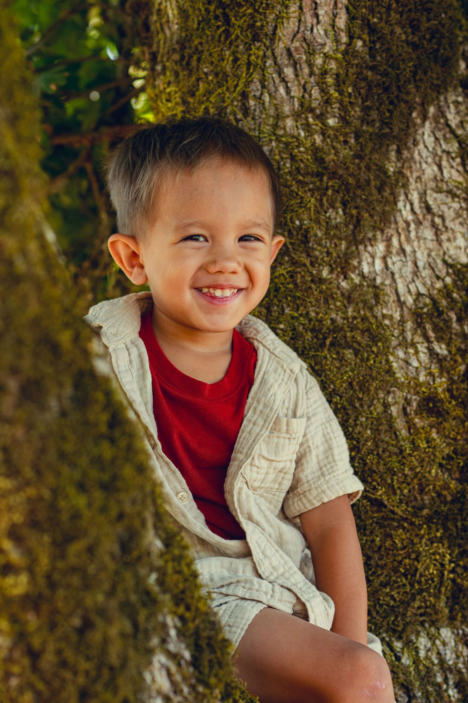 A child in a red shirt and beige vest poses playfully against a mossy tree trunk in an outdoor setting.
