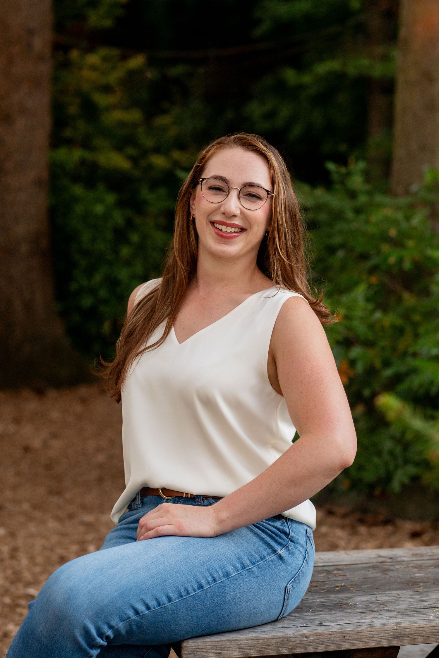 A person in a white sleeveless top and blue jeans sits casually on a wooden surface with trees in the background.