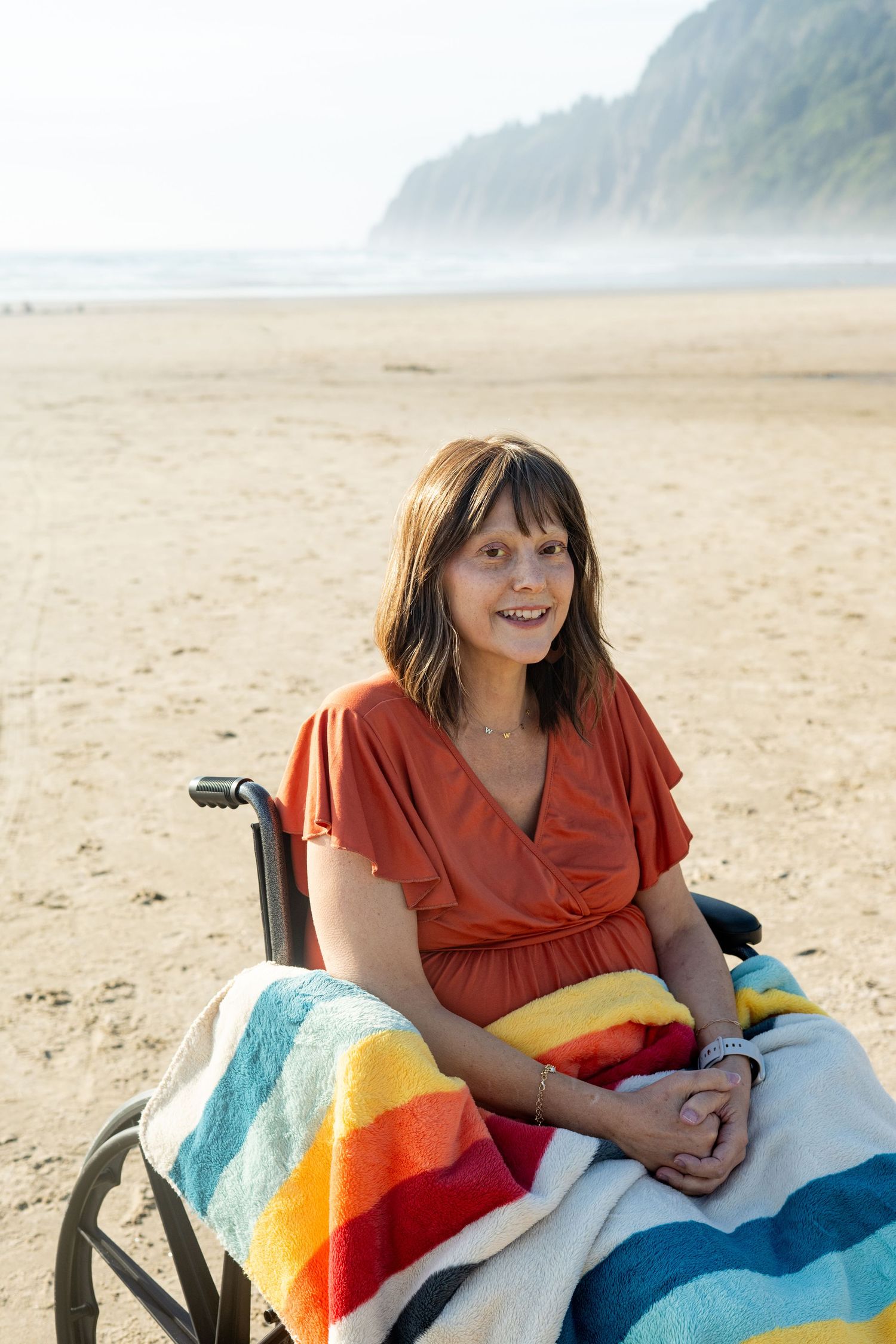 A person wrapped in a colorful striped beach blanket smiles while sitting in a wheelchair on a misty beach.