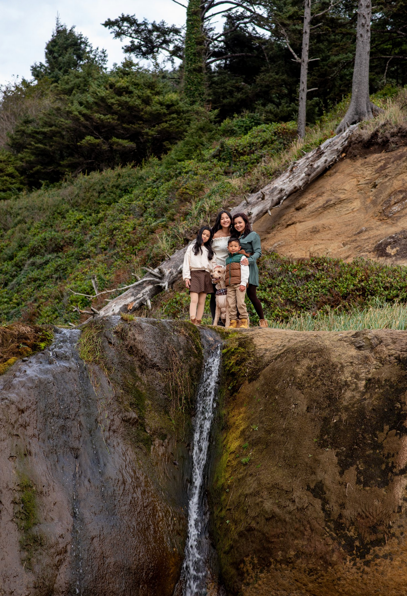 A group poses together on a cliff edge with a small waterfall and forest backdrop.