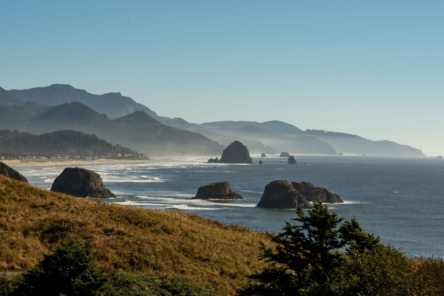 Panoramic view of the Oregon coast with waves crashing against rugged rock formations and misty mountains in the distance.