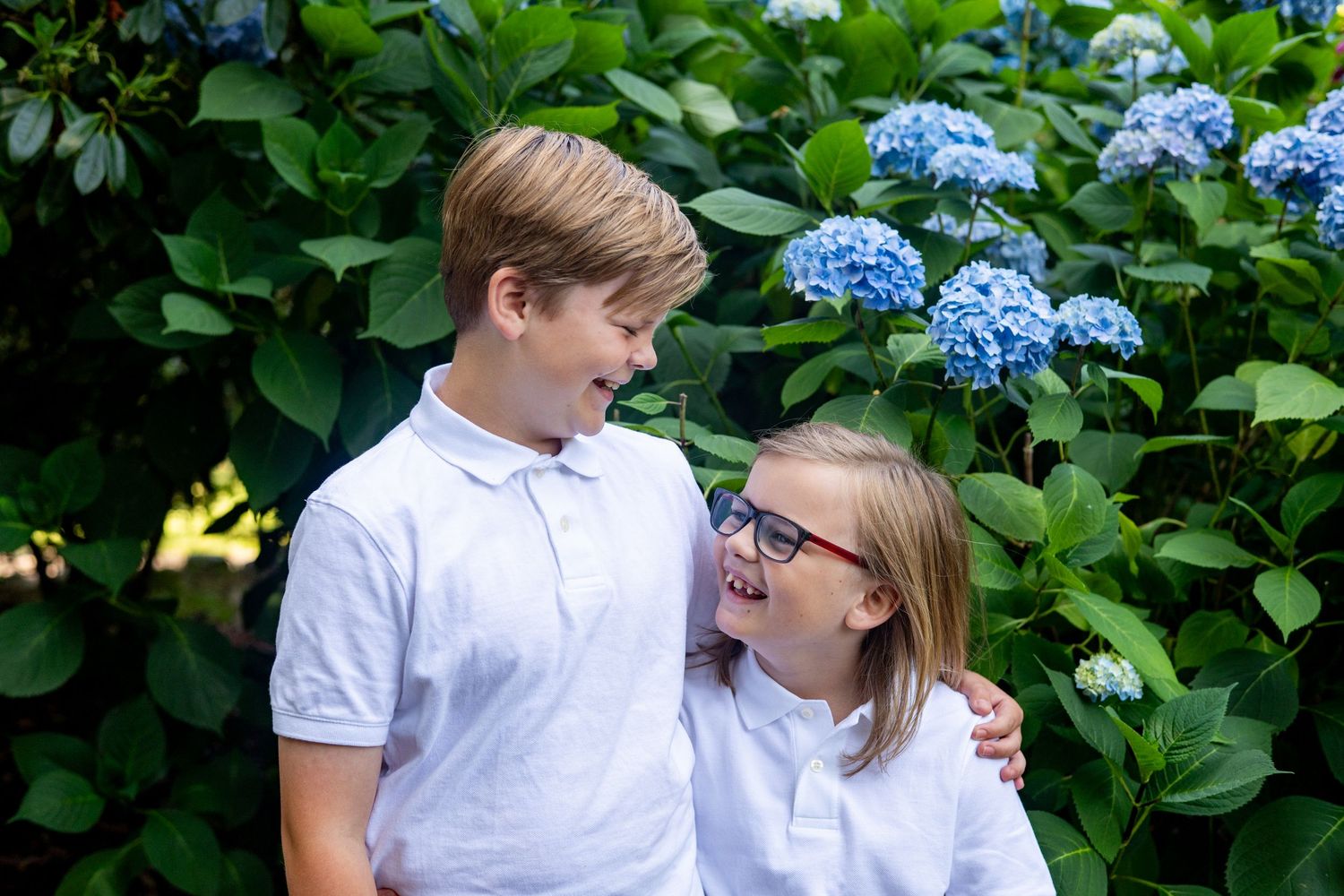 Two young children in white polo shirts stand together smiling in front of blooming blue hydrangea bushes.