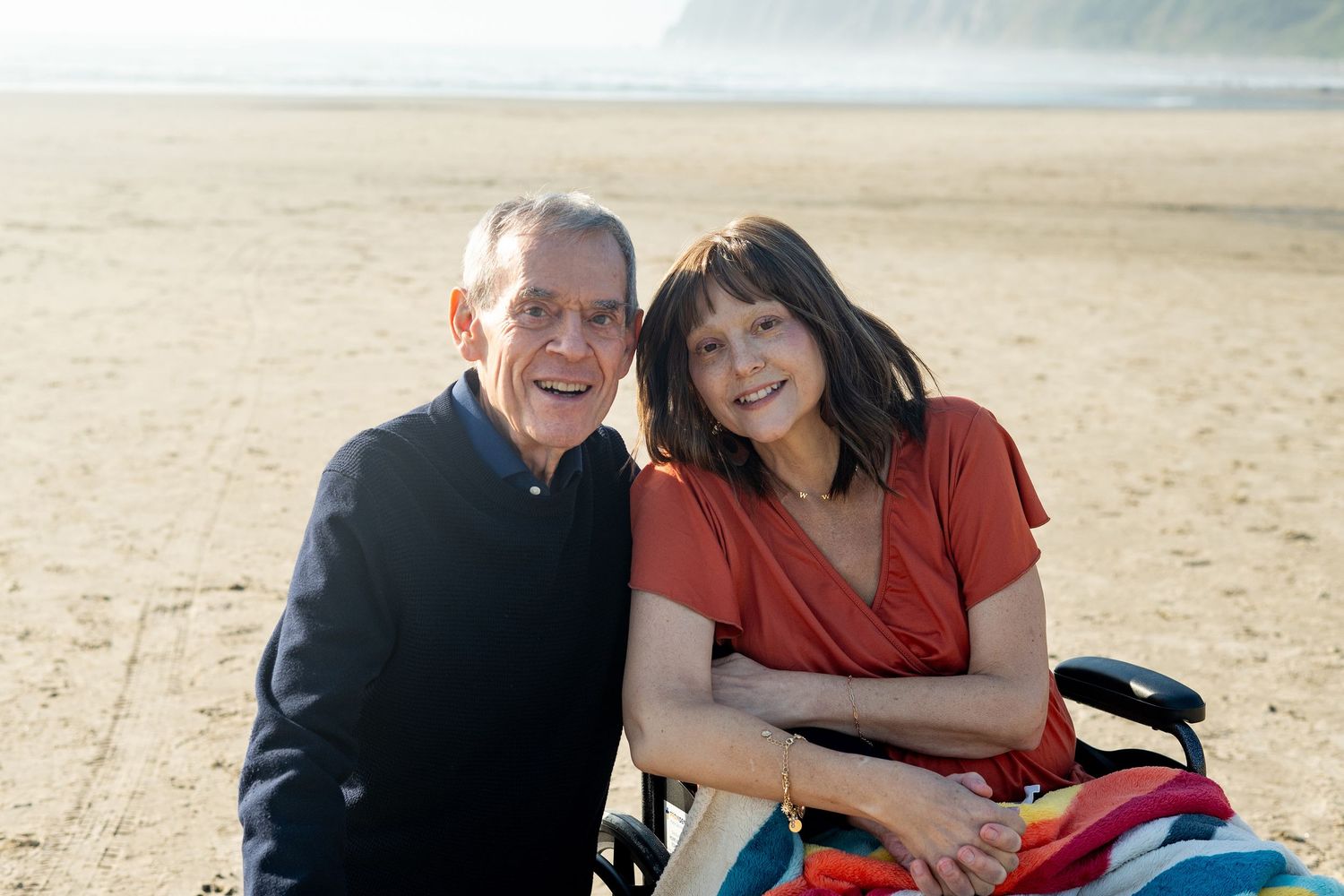 Two people laughing and embracing while having a playful moment on a sandy beach near ocean waves.