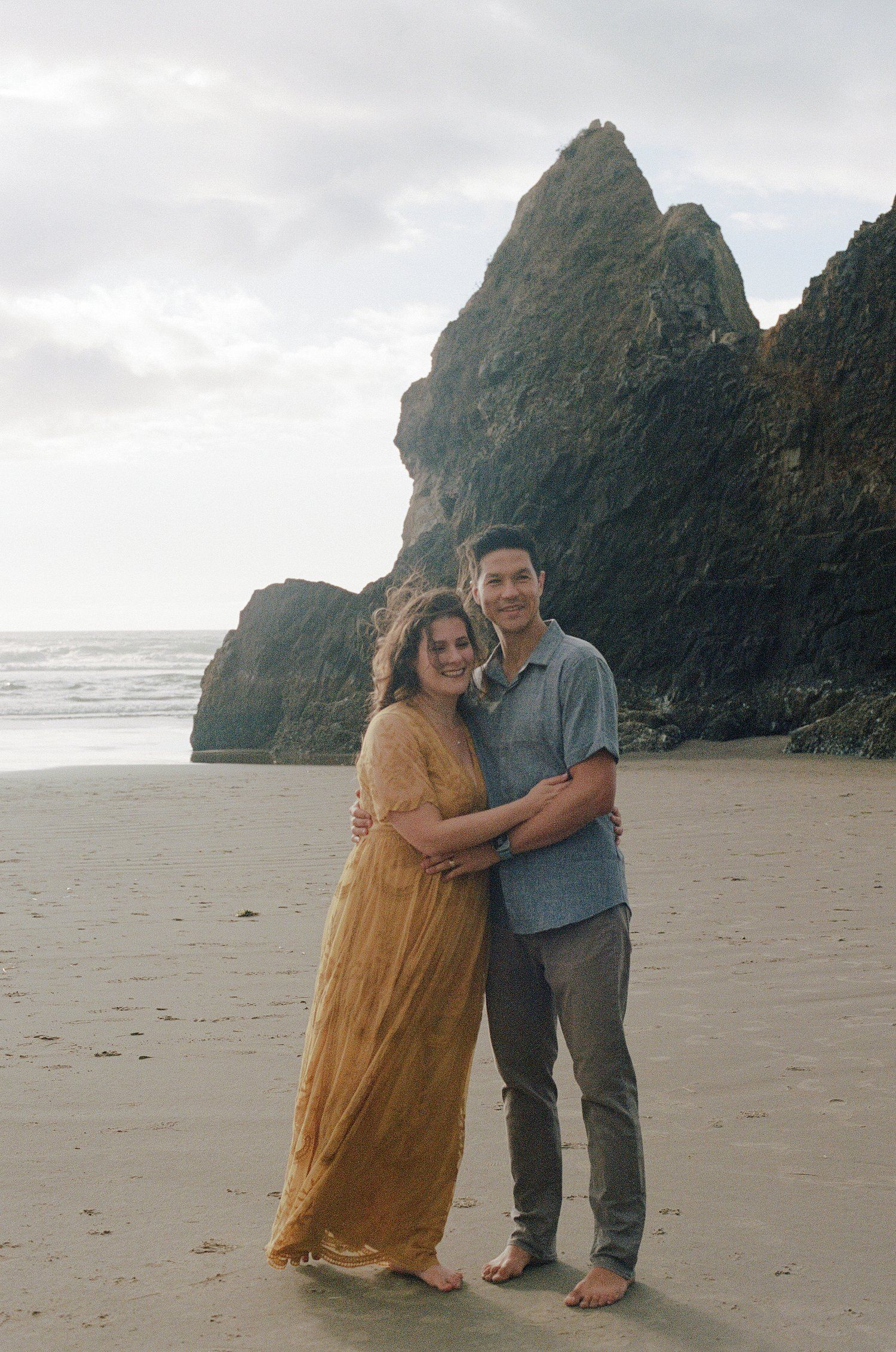 Couple poses together on sandy beach with dramatic rock formations and ocean waves in background.