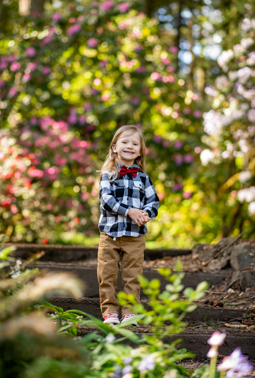 A child in plaid shirt and brown pants stands on a nature trail surrounded by pink flowers and greenery.