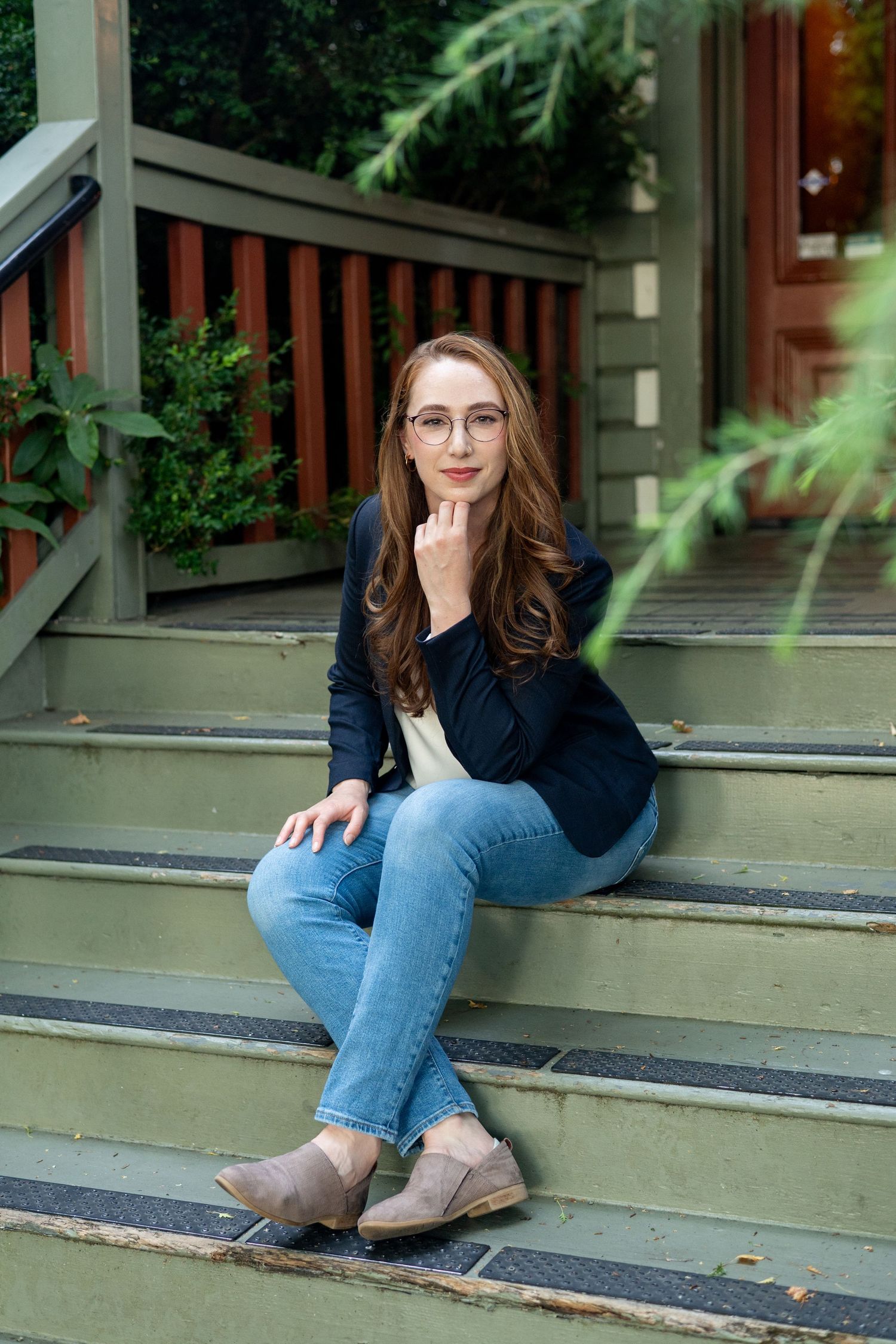 A person in a navy blazer and jeans sits casually on green-painted wooden steps outside a house with greenery.