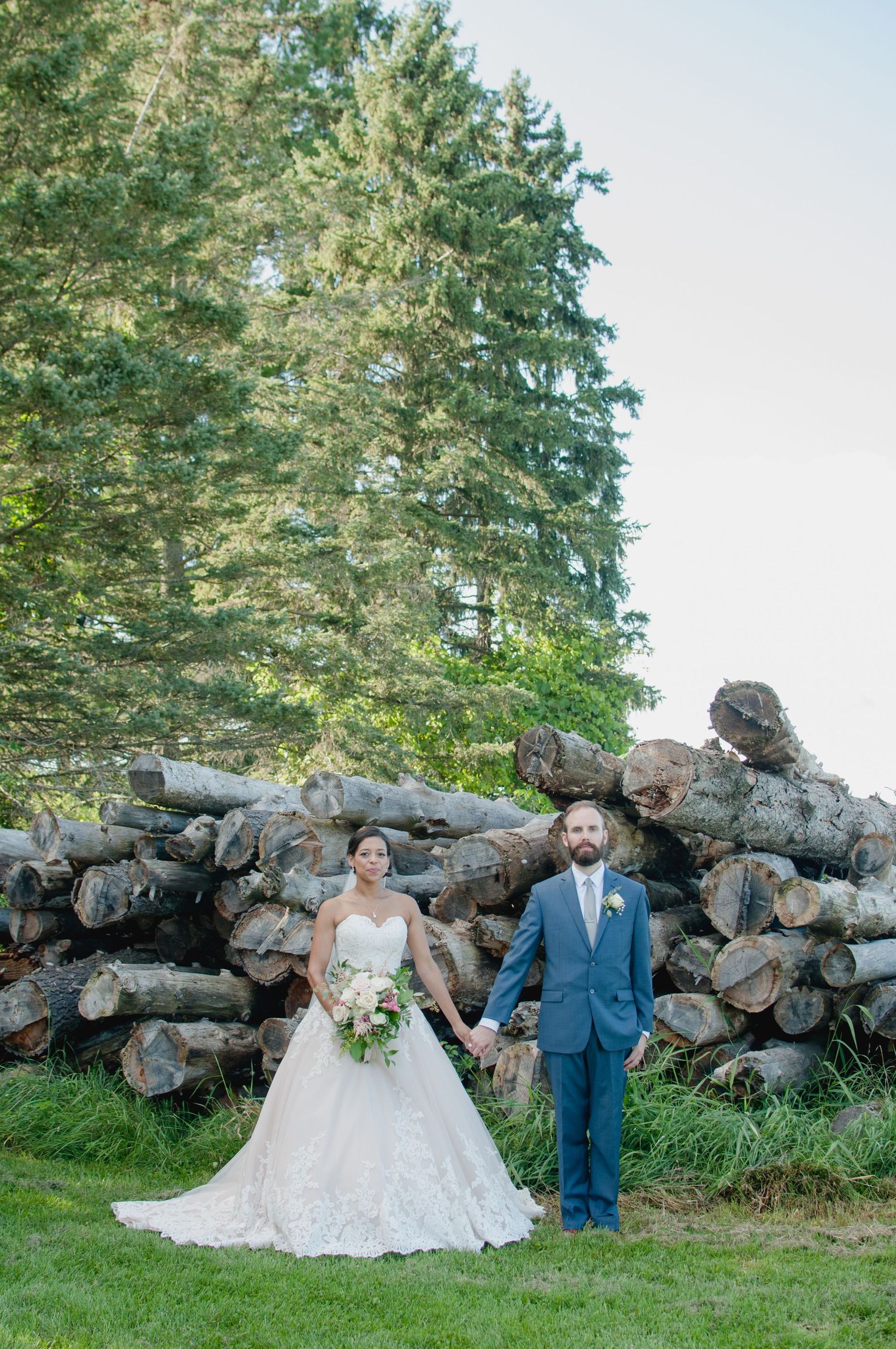 A newlywed couple poses against a rustic log wall backdrop with evergreen trees.