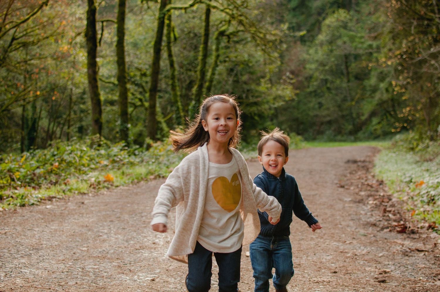Two children running joyfully down a wooded trail surrounded by autumn foliage.
