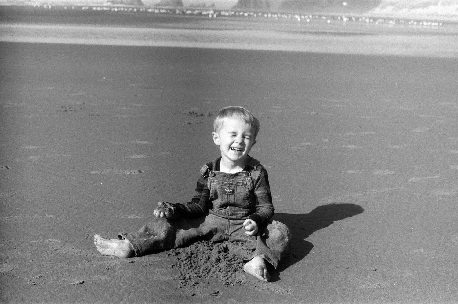 A black and white vintage photograph shows a small child sitting and playing on a sandy beach.