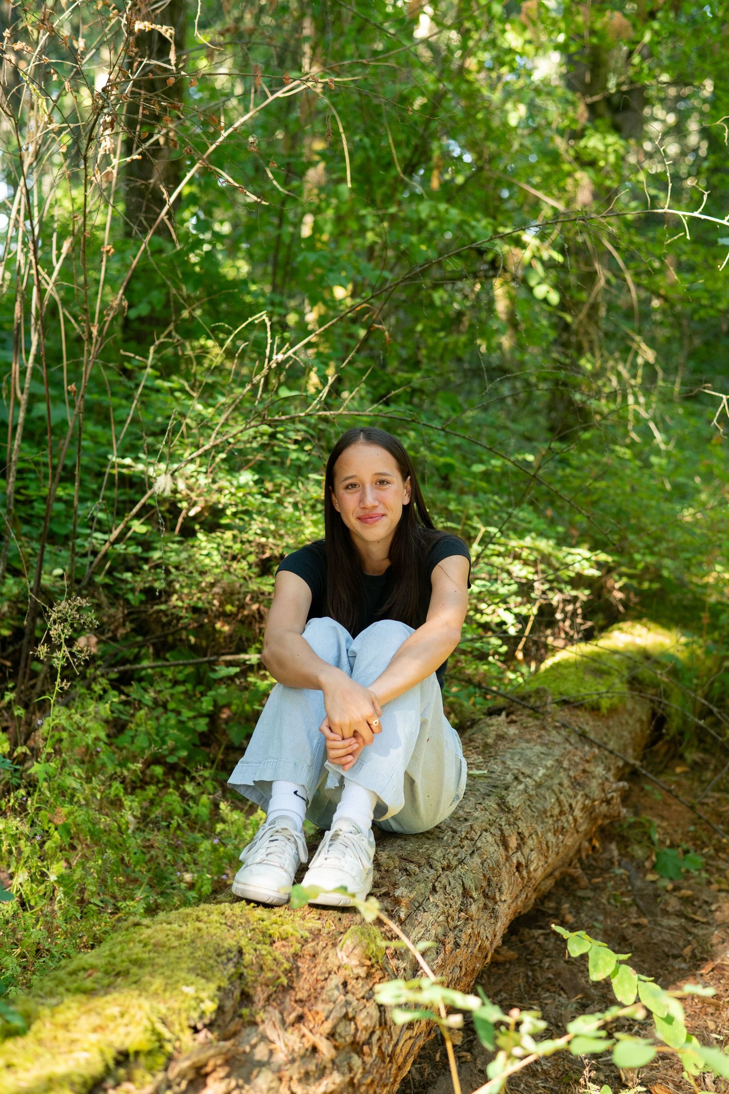 A person in a black shirt and light pants sits relaxed on a moss-covered log in a lush green forest setting.