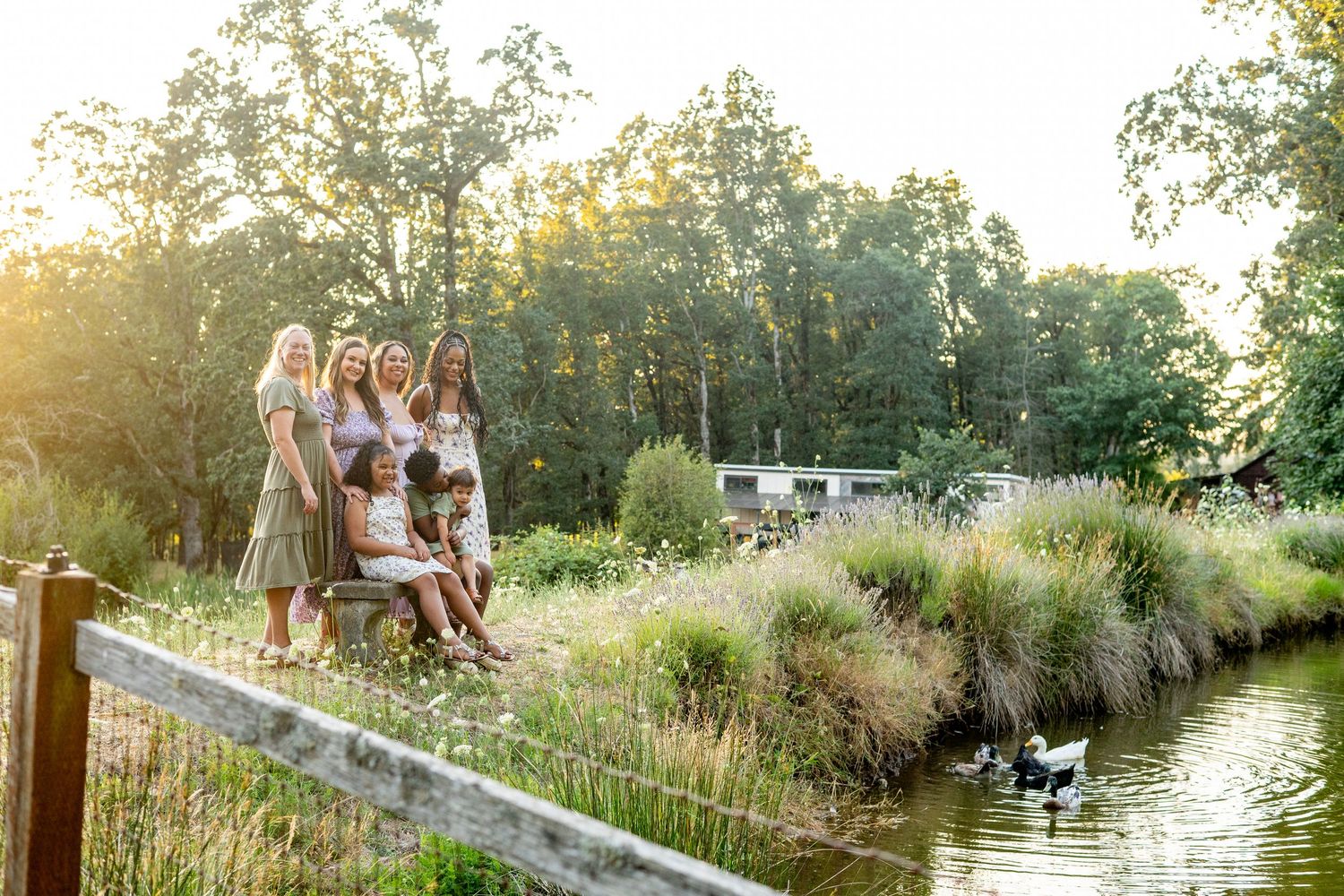 A group sits together on a wooden dock overlooking a pond during golden hour while their dog plays in the water below.
