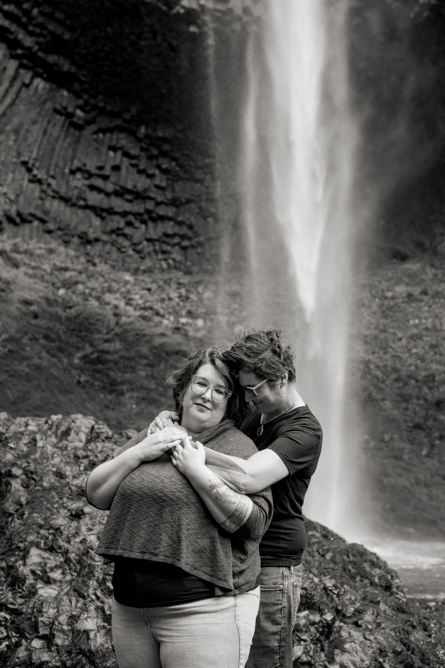 A romantic embrace captured in black and white against a scenic waterfall backdrop in nature.