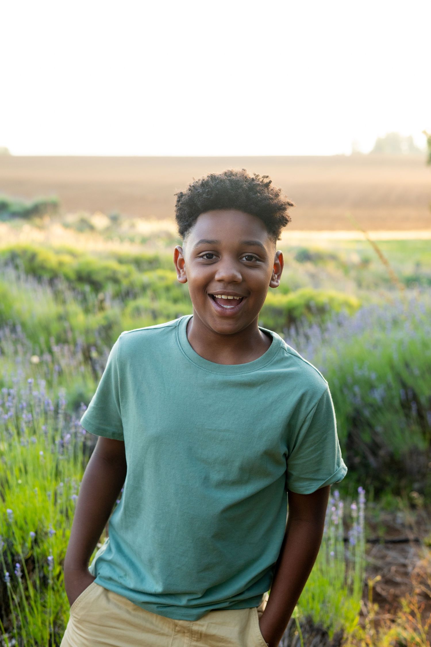 A child in a teal t-shirt smiles joyfully while sitting outdoors in a field of purple lavender flowers at sunset.