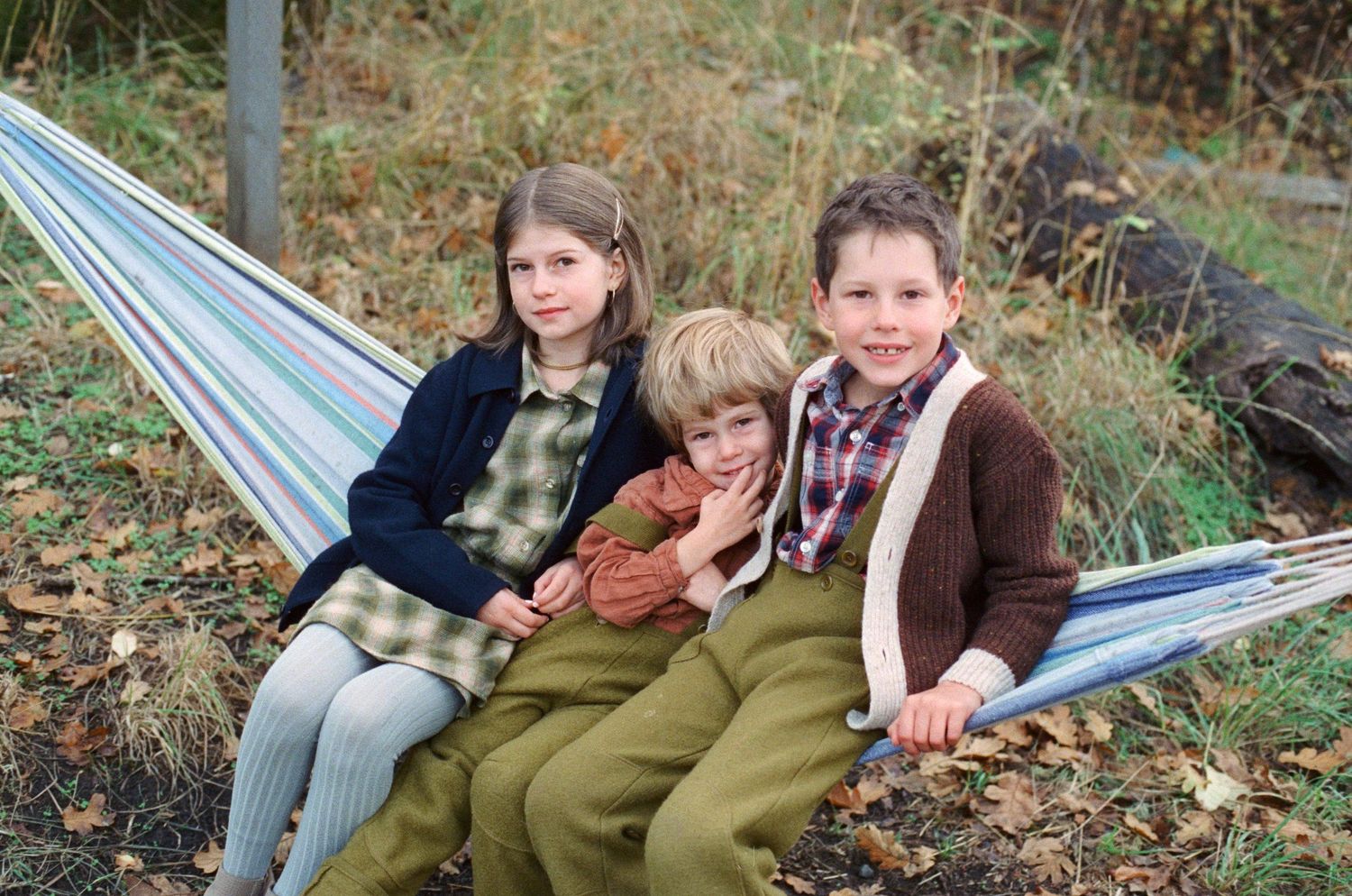 Three people sit together on a striped hammock outdoors in autumn.