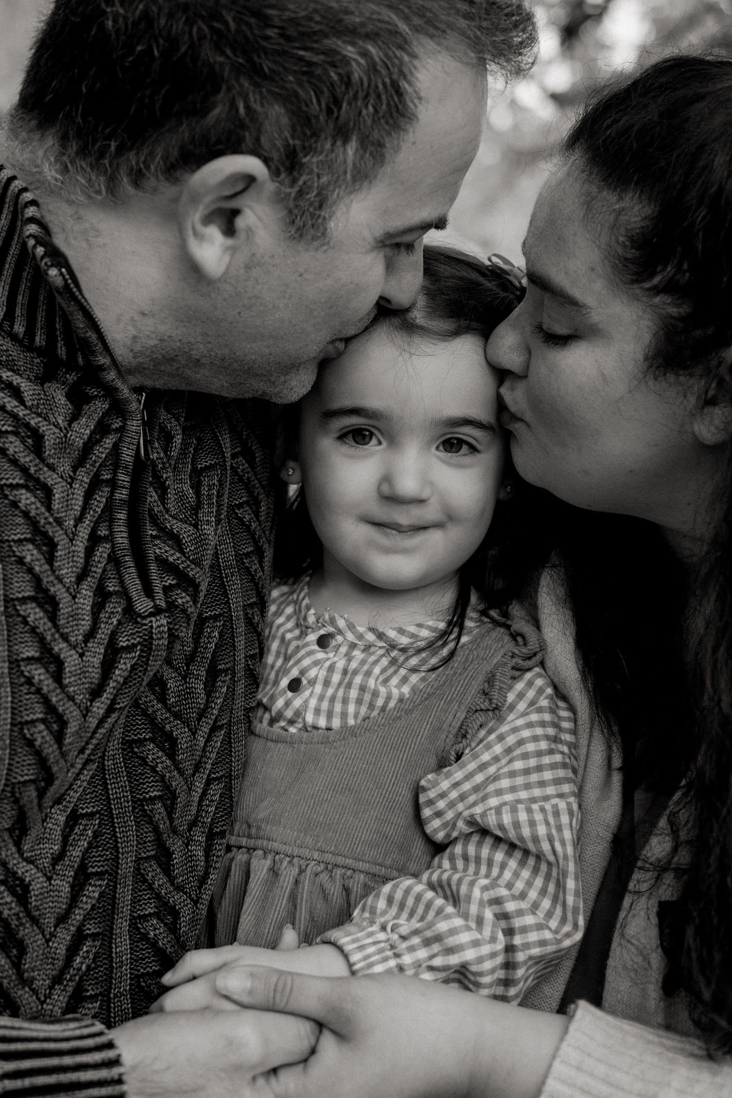 A heartwarming black and white portrait showing parents tenderly kissing their young child in a gingham dress.