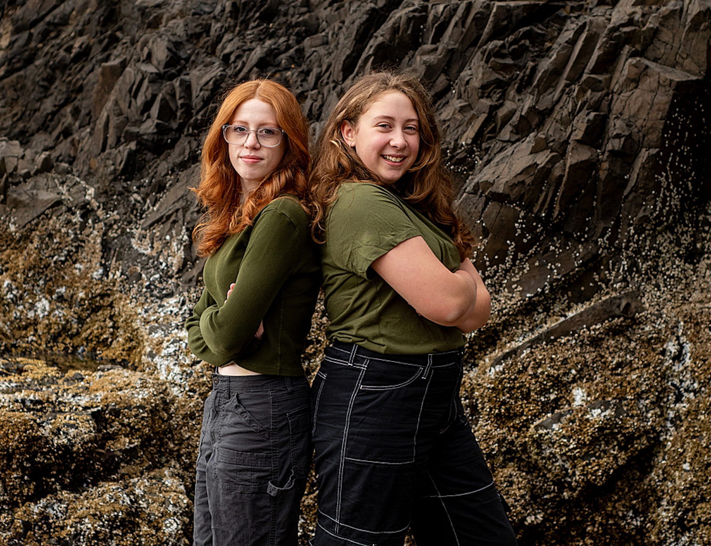 Two sister pose back-to-back against textured rocky coastal cliff background.