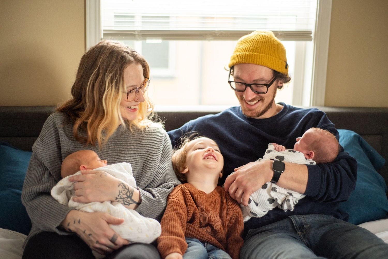 Family relaxing together on a couch by a sunny window with small children.