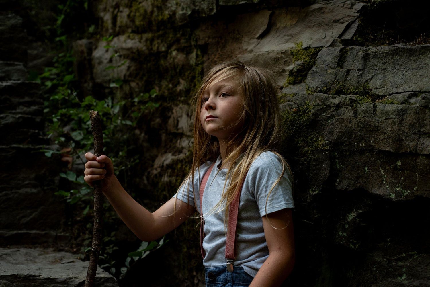 A young explorer in suspenders and t-shirt stands against a rocky wall in moody natural lighting.