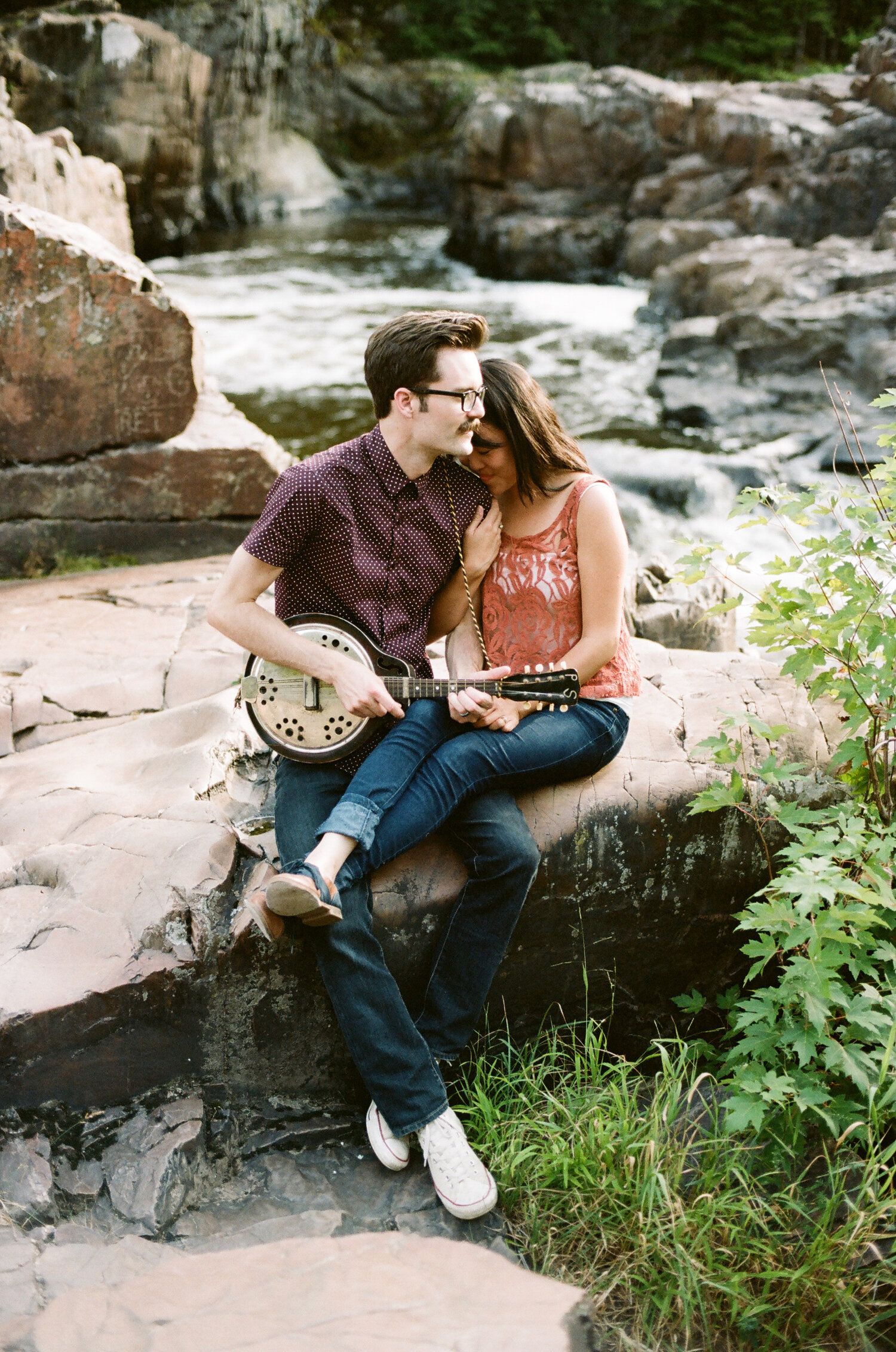 A couple sitting on rocky ledge by a stream share a tender moment with a musical instrument.