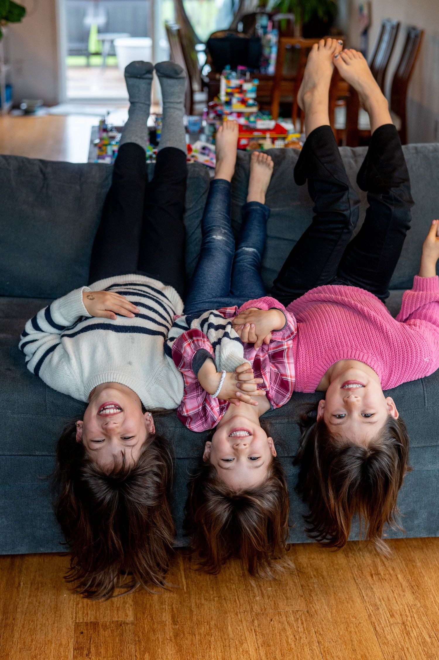 Three siblings laying upside down on a couch with their hair hanging down and feet up in a playful moment.