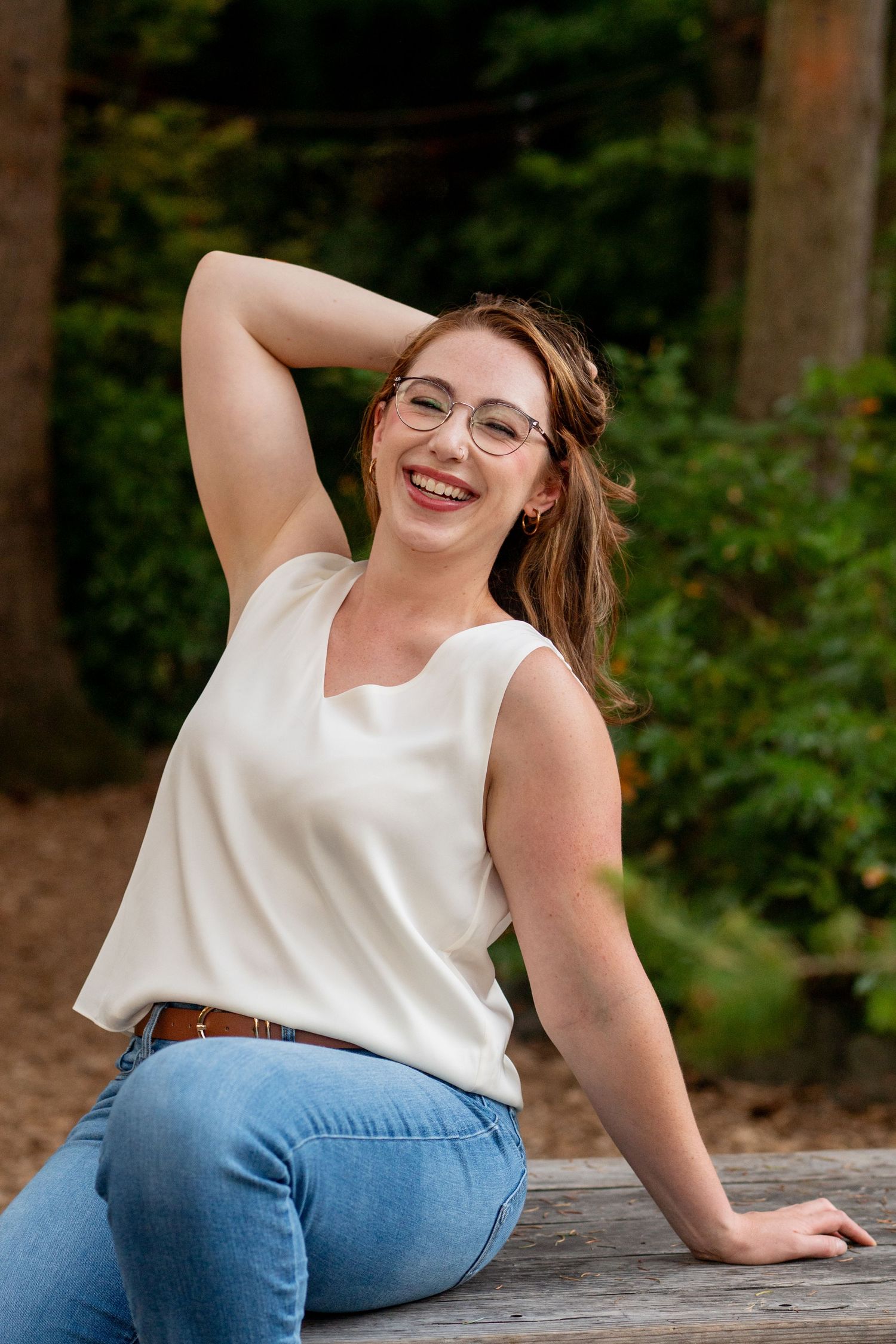 Woman in white sleeveless top and jeans sits casually on wooden surface outdoors with greenery in background.