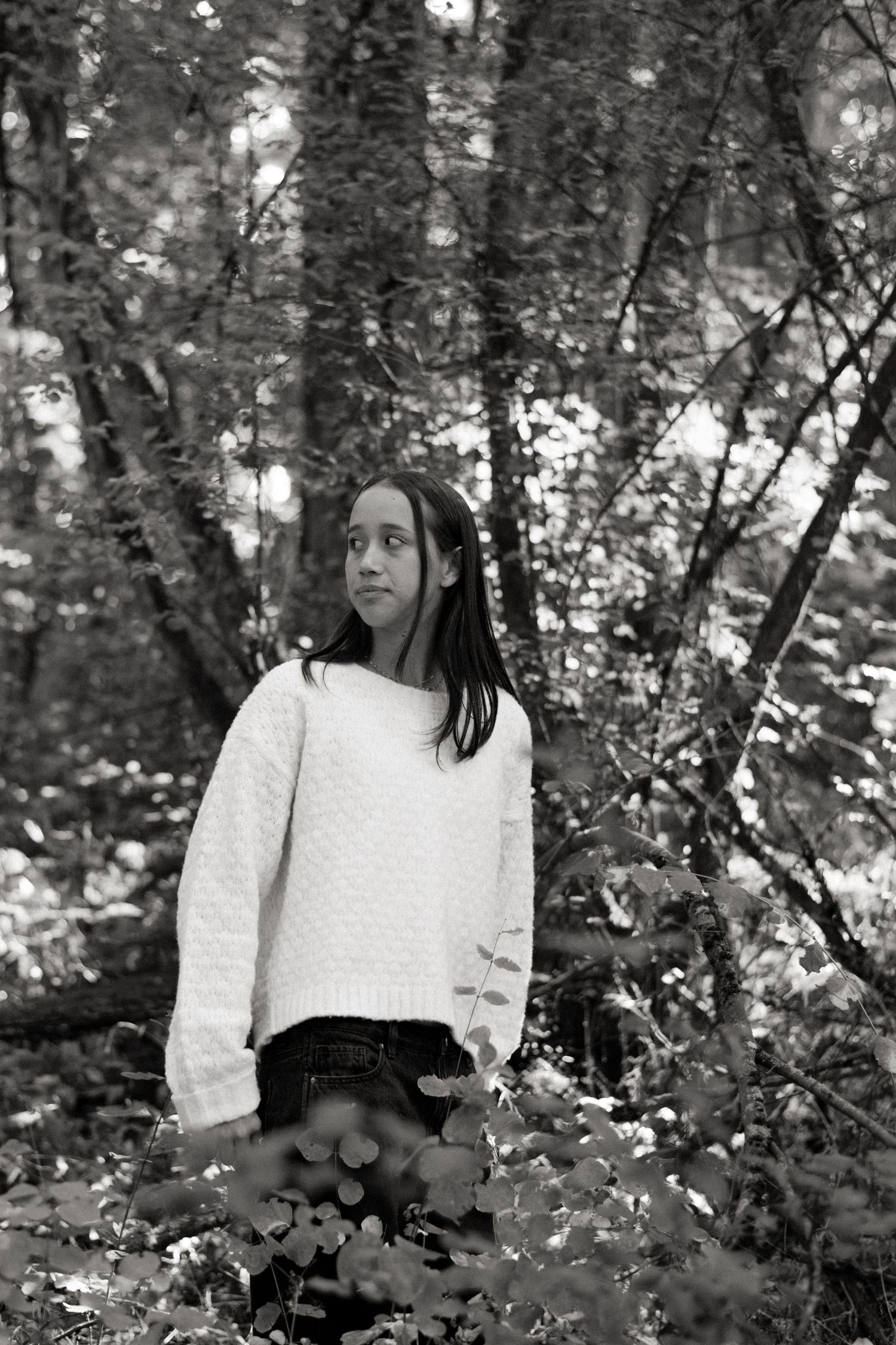 A figure in a white sweater stands among trees in a black and white forest scene.