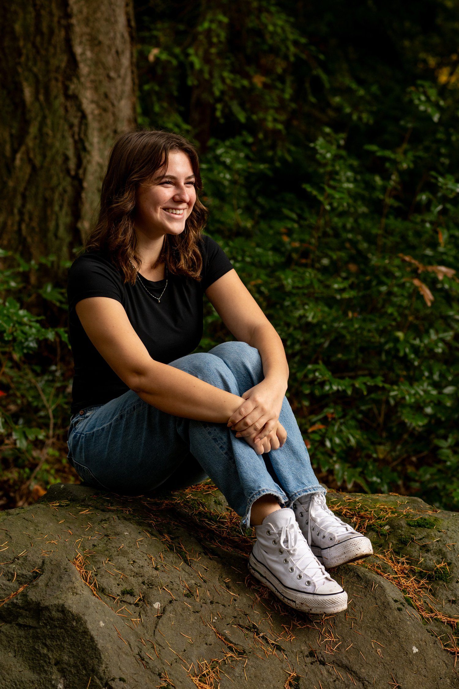 A casual portrait on a rock wearing jeans and white sneakers in a forest setting.