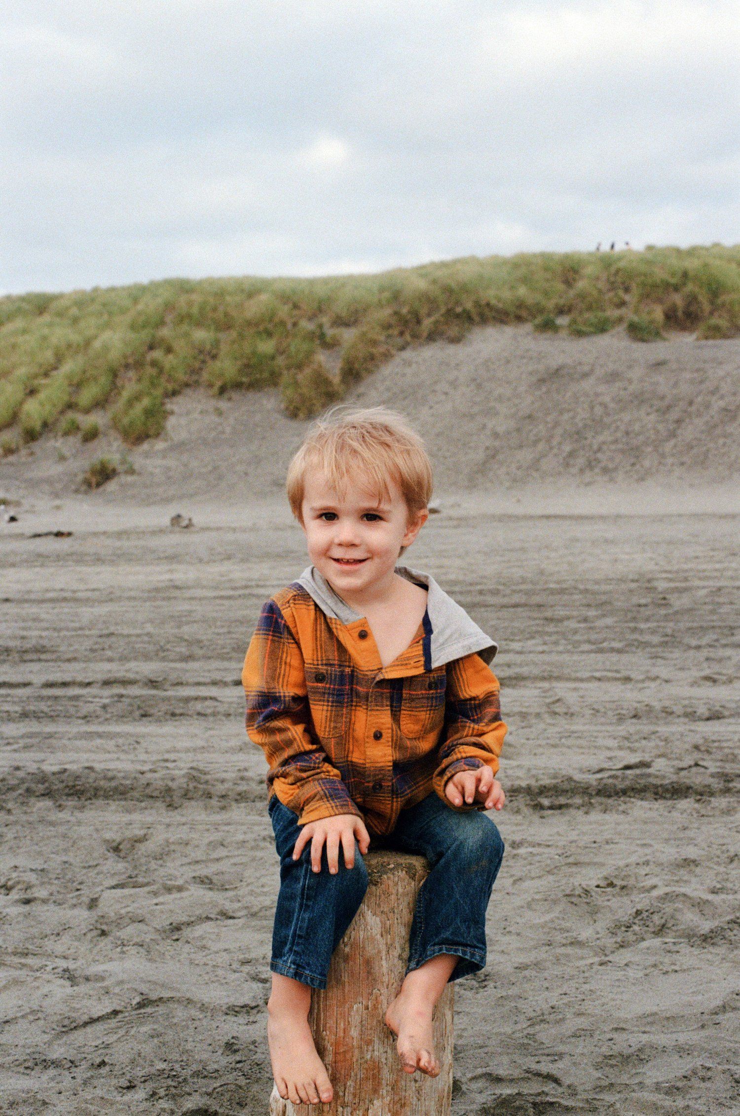 A child in orange jacket sits barefoot on wooden log at beach.