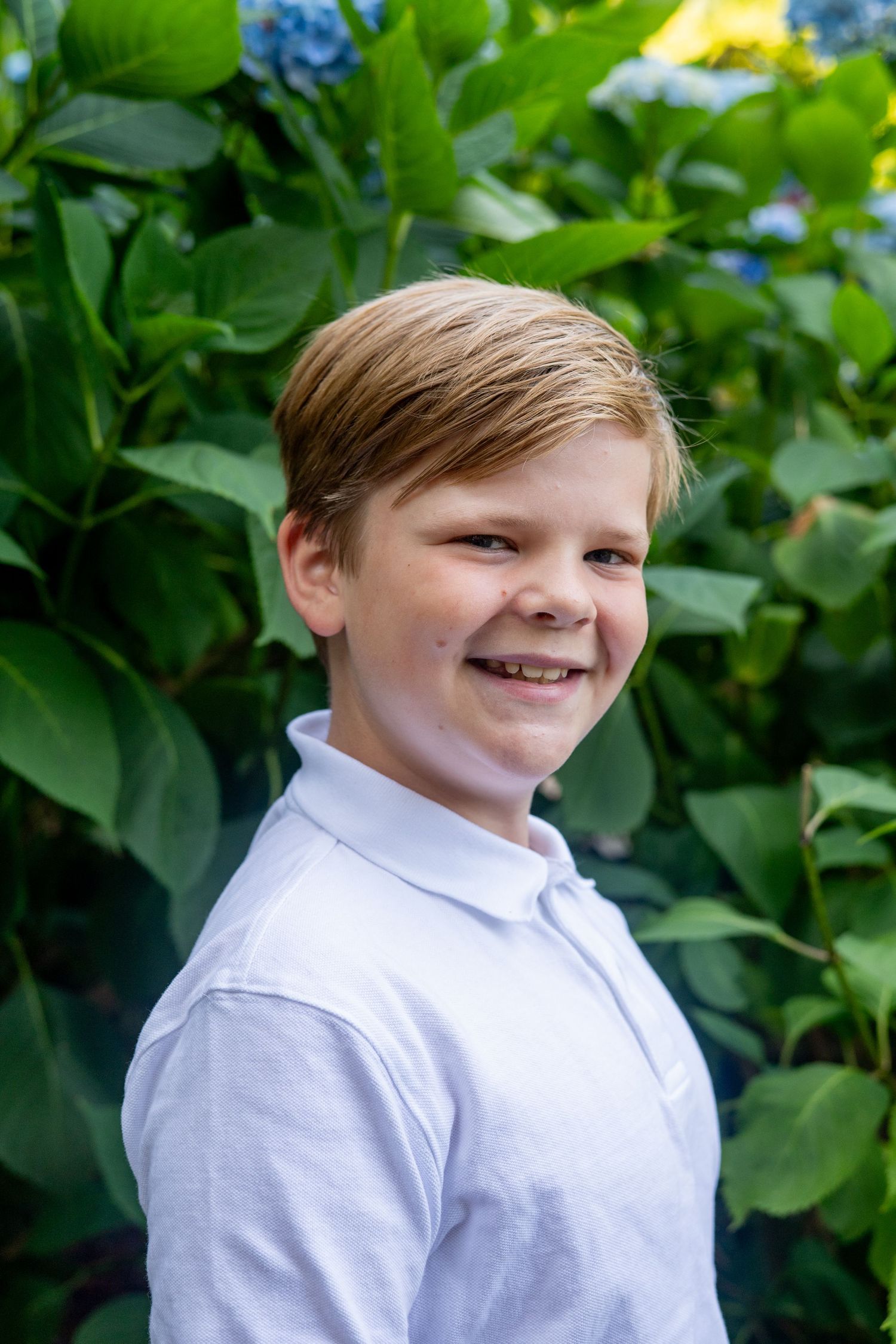 A young person wearing a white collared shirt smiles outdoors against a background of green leaves and blue flowers.
