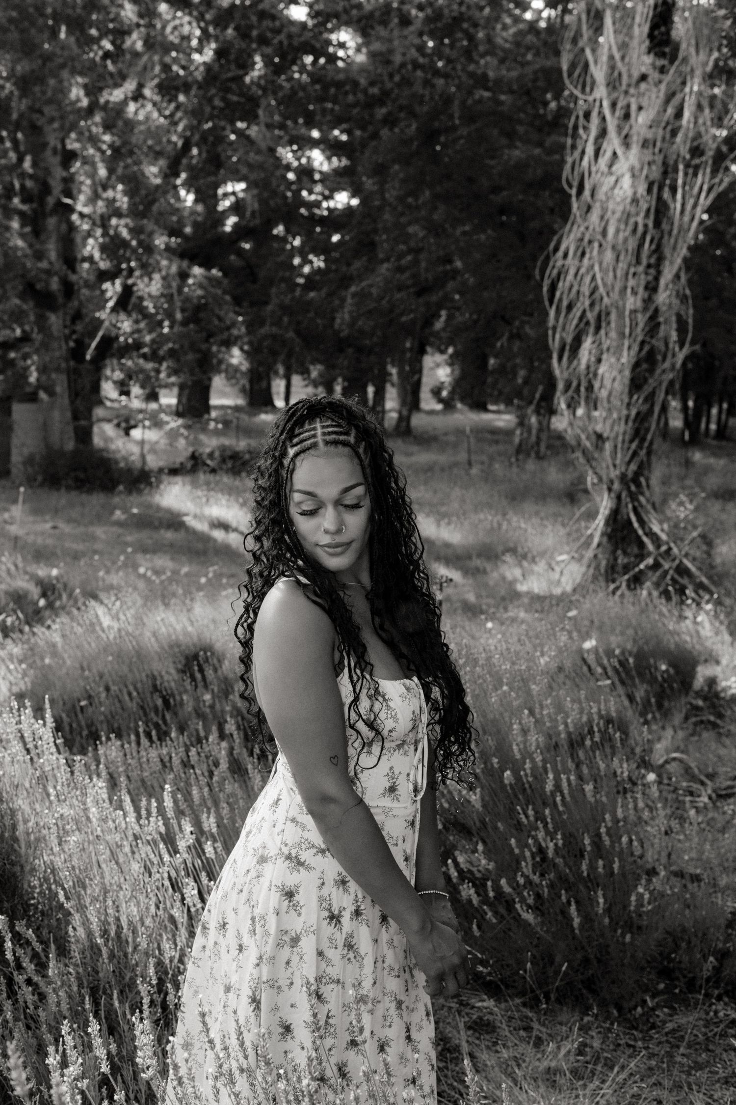 Black and white portrait of someone in a floral dress standing in a dreamy outdoor setting with tall trees and lavender.