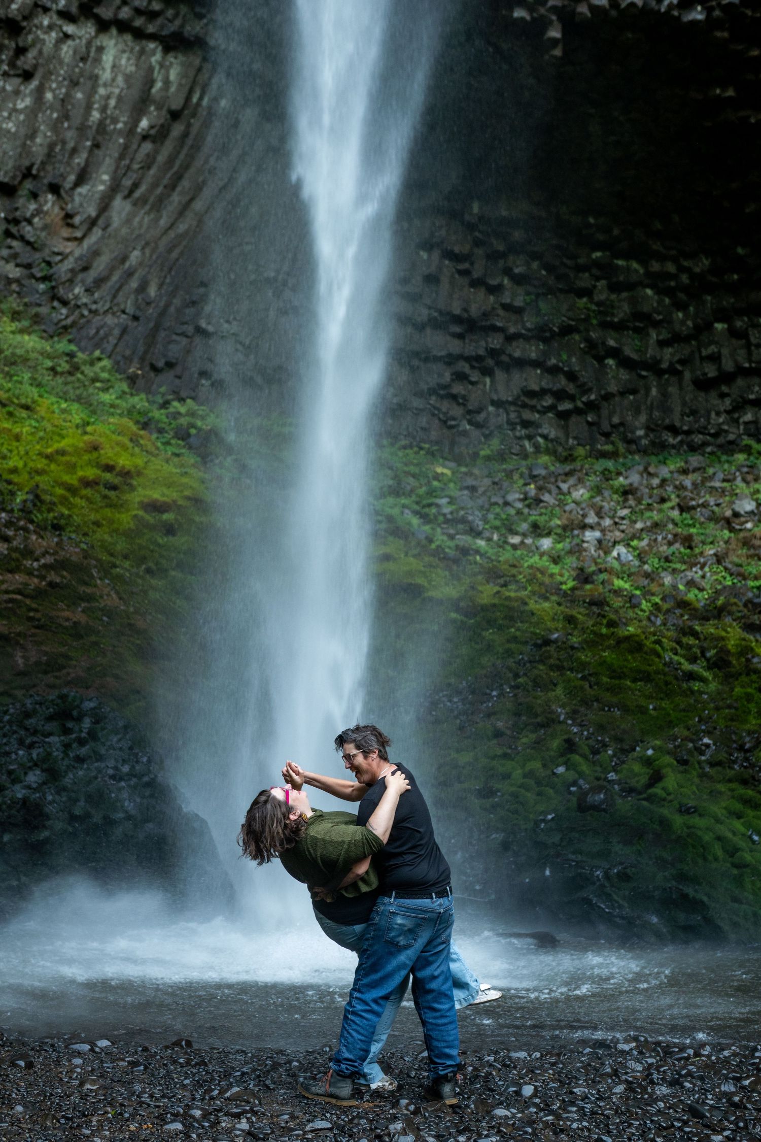 Couple poses playfully in front of a majestic waterfall cascading down moss-covered cliffs.