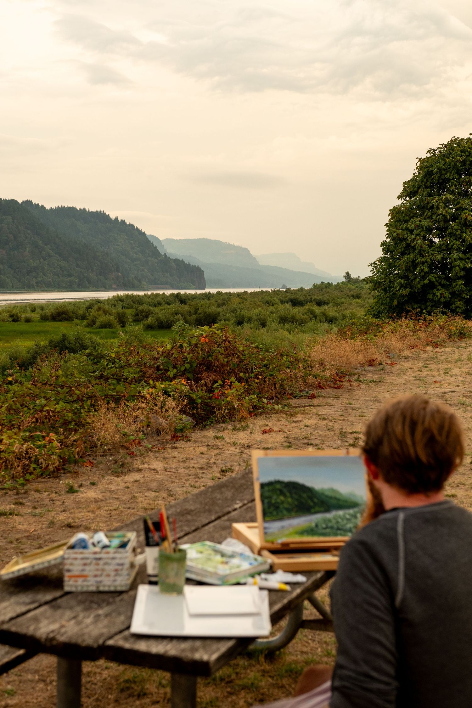 Person sits sketching at outdoor table overlooking misty mountain landscape and waterfront view.