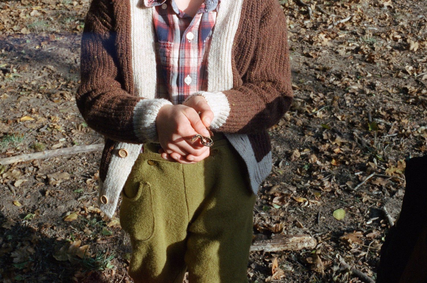 Close-up of clasped hands wearing a brown cardigan and plaid shirt against an outdoor background.